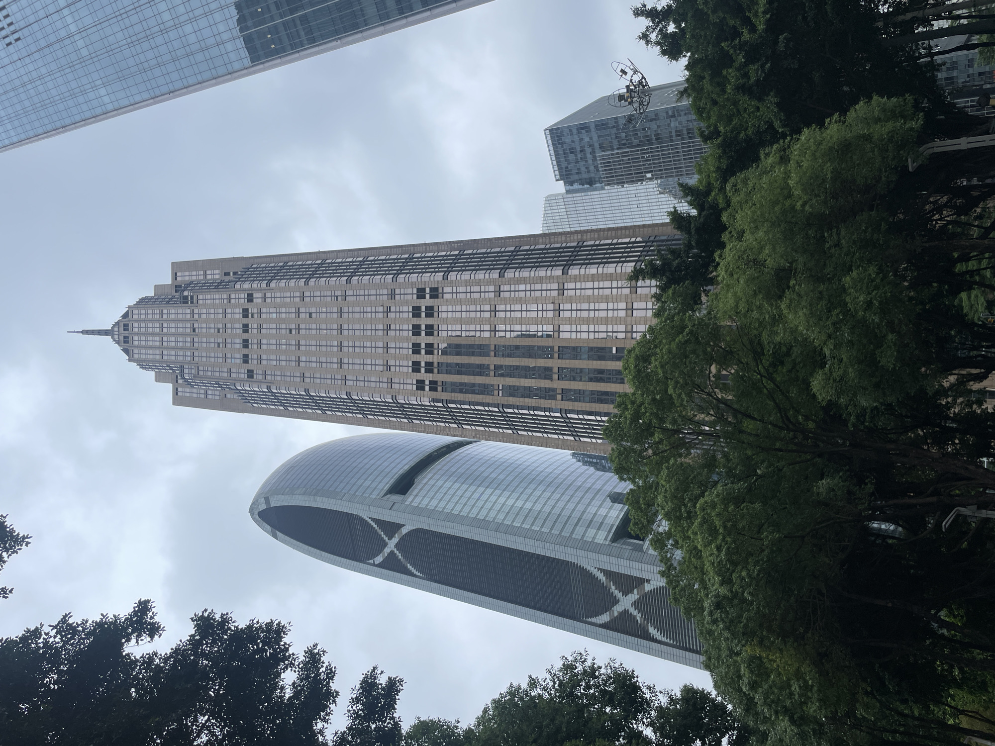 The image captures a view looking upwards at several skyscrapers in Tianhe District, Guangzhou, China. The most prominent building is the Guangzhou CTF Finance Centre, a tall skyscraper with a beige exterior and a spire at the top. Adjacent to it is a rounded skyscraper with an "X" pattern along its sides. Portions of other skyscrapers are visible at the top of the frame. Trees with green foliage occupy the bottom of the image, partly obscuring the lower portions of the buildings. The sky is overcast, suggesting a cloudy day. No people are visible in the frame.