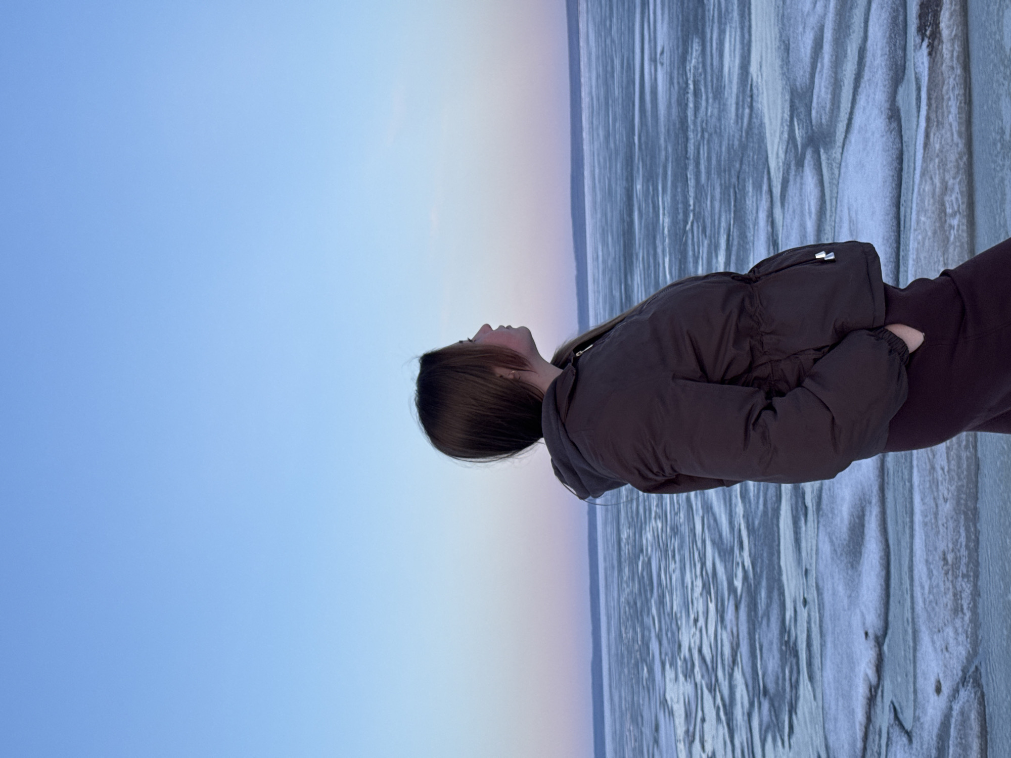 A girl in a brown puffer jacket is standing on what appears to be a frozen body of water. The background shows a clear blue sky fading to a pink hue near the horizon. The body of water has a rough, textured surface with visible cracks and patterns in the ice. The location is Noviy, Russia.
