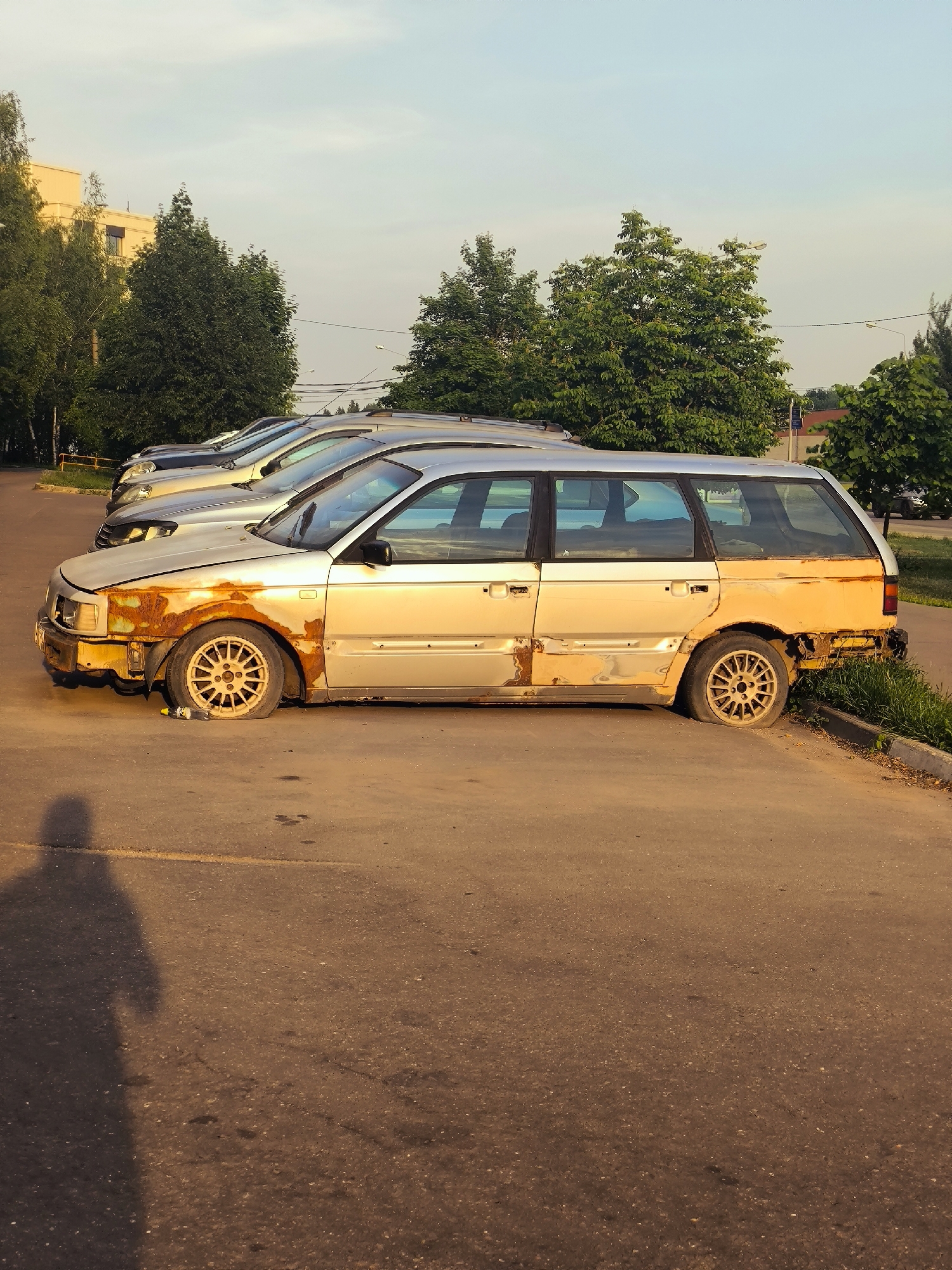 The image depicts a side view of a heavily rusted, older model station wagon parked in an outdoor lot. The car is silver with significant patches of orange rust, particularly on the front fender and along the lower body panels. One of its front tires appears to be flat, and a plastic bottle is visible on the ground near it. The car is parked behind other similar vehicles, suggesting a lineup in a parking area.
The setting is an asphalt lot, possibly a parking lot, with a grassy verge and some vegetation visible on the right side. In the background, there are trees and a building that appears to be a light-colored, multi-story structure. The sky above is a pale blue, indicating daytime. The lighting suggests it might be late afternoon or early morning, with a warm, golden hue casting a shadow of an unseen person onto the asphalt in the foreground. The overall mood conveyed by the image is one of decay and abandonment, primarily due to the condition of the car. There is no visible text in the image.