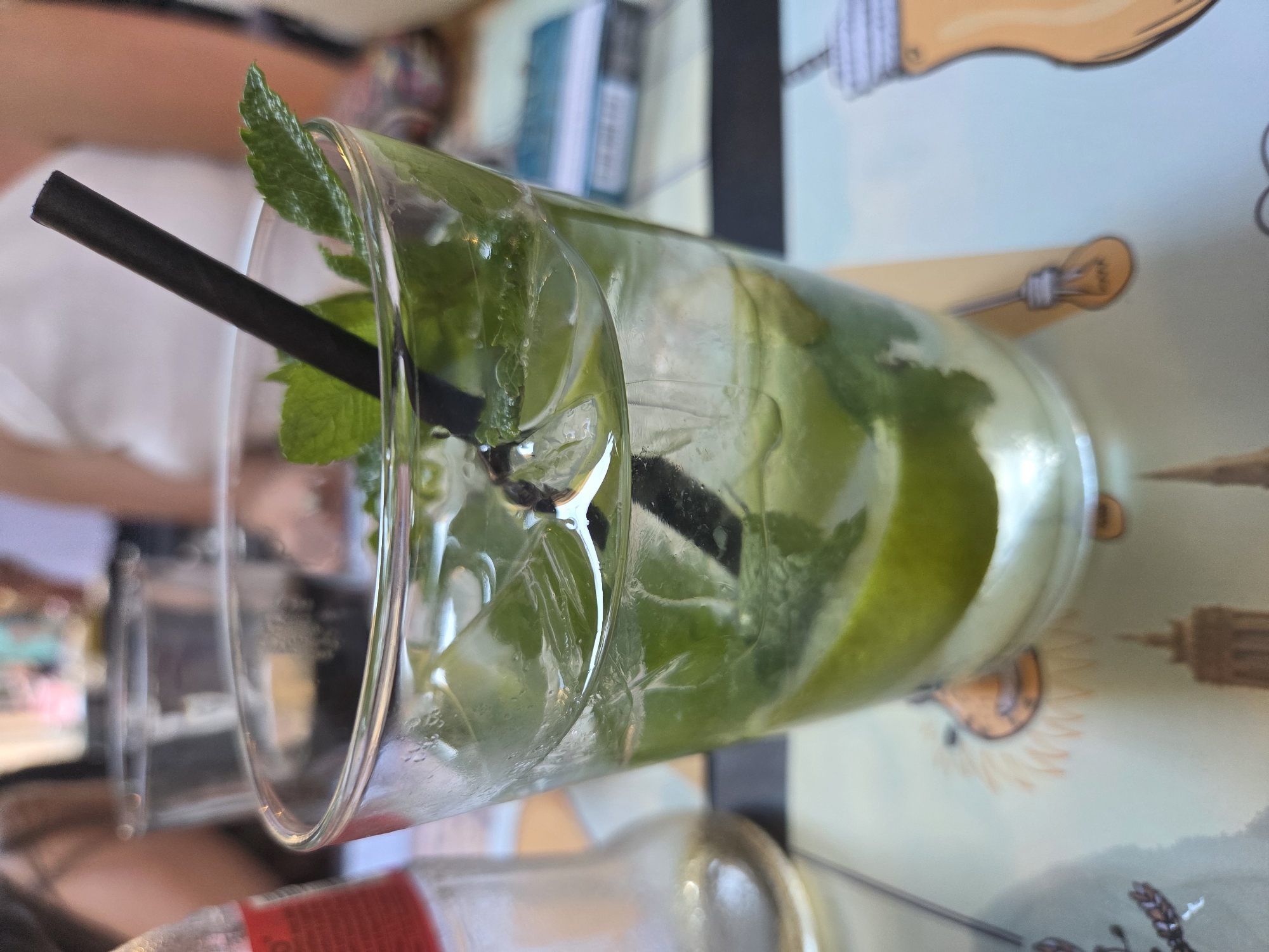 A tall glass of Mojito sits on a patterned table. The Mojito contains ice cubes, mint leaves, and lime slices, and a black straw is visible. The table has a light-colored background with illustrations of light bulbs and city buildings, including what appears to be a tower resembling the Eiffel Tower. Parts of two people are visible; one is wearing a white shirt.