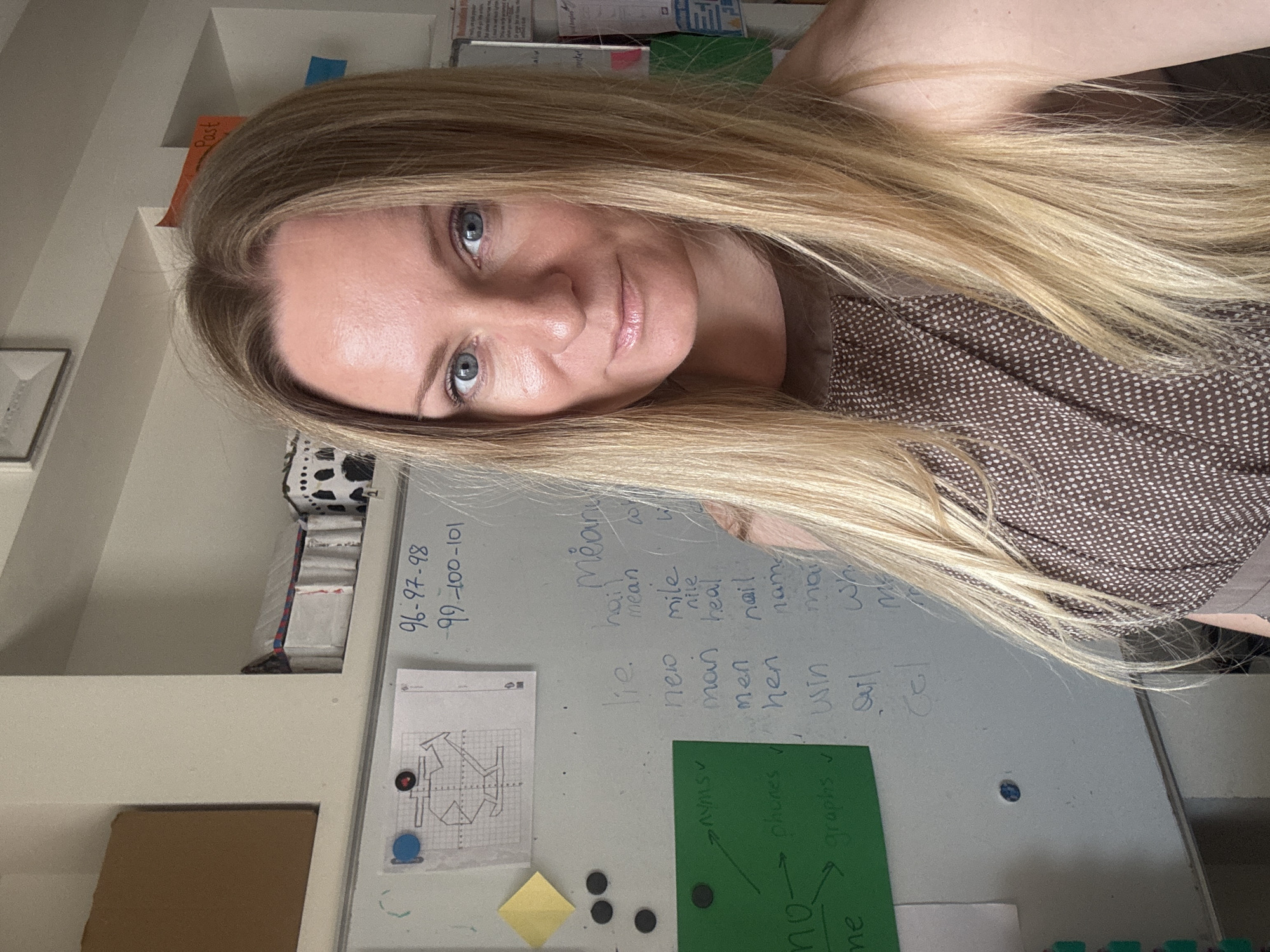 A woman with blonde hair is standing in front of a whiteboard that has writing and drawings on it. She is looking at the camera and smiling. The whiteboard has a drawing of a complex geometric shape at the top, and below it, lists of words and numbers. To the left of the whiteboard, there are several sticky notes and magnets. The woman is wearing a brown top with a patterned design. The room appears to be an indoor space, possibly a classroom or office, with shelves and boxes visible in the background. There is no indication of the city of Budva, Montenegro in the image.