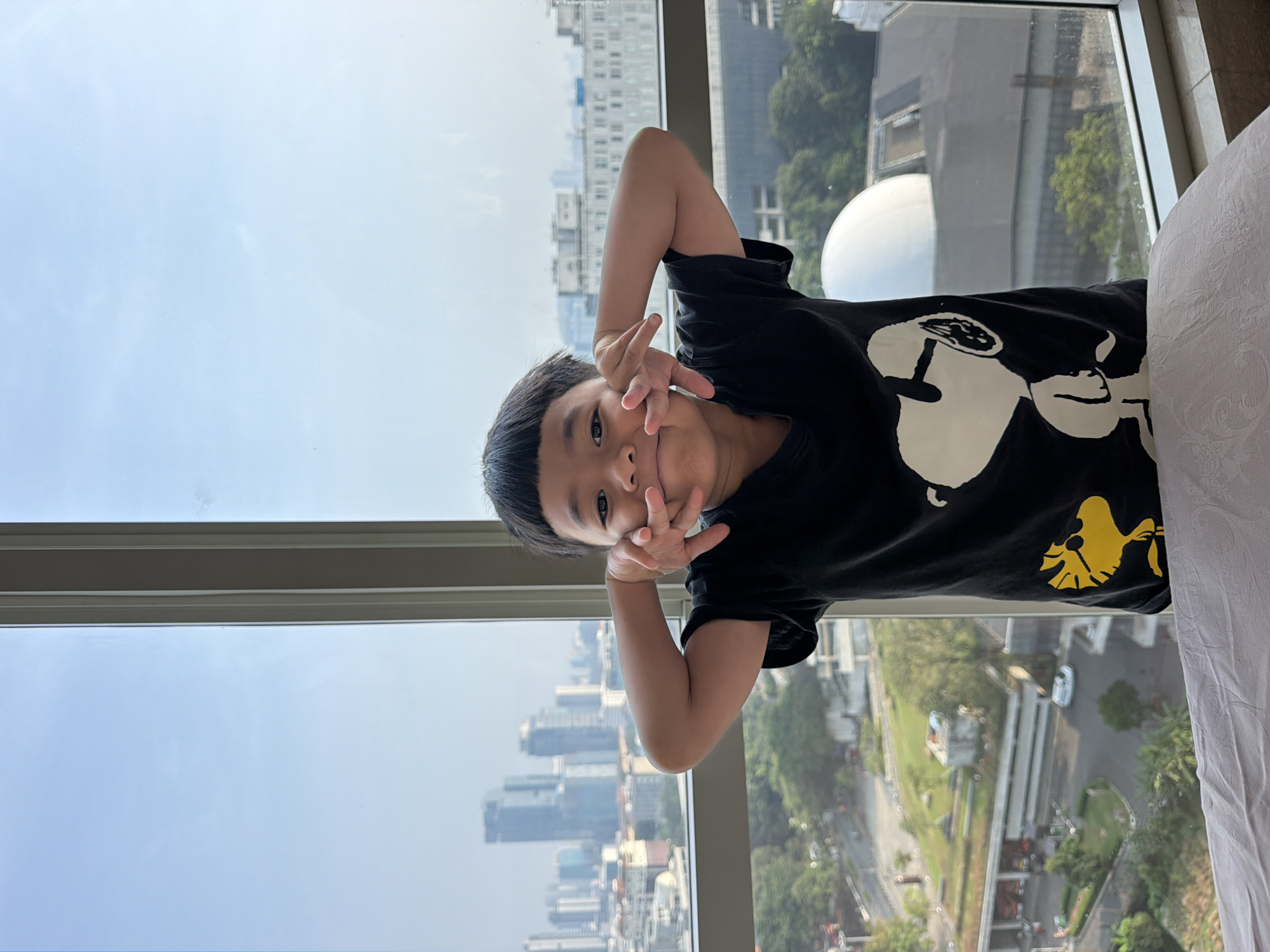 A young boy, likely of Southeast Asian descent, stands in front of a large window overlooking a city skyline. He is wearing a black t-shirt with a Snoopy graphic. The boy is making a playful gesture with his hands near his face, with his fingers spread. The window offers a view of tall buildings, trees, and a street below, suggesting an urban setting in Central Jakarta, Indonesia. The sky is clear and blue. The boy is the central subject of the image, positioned in the foreground.