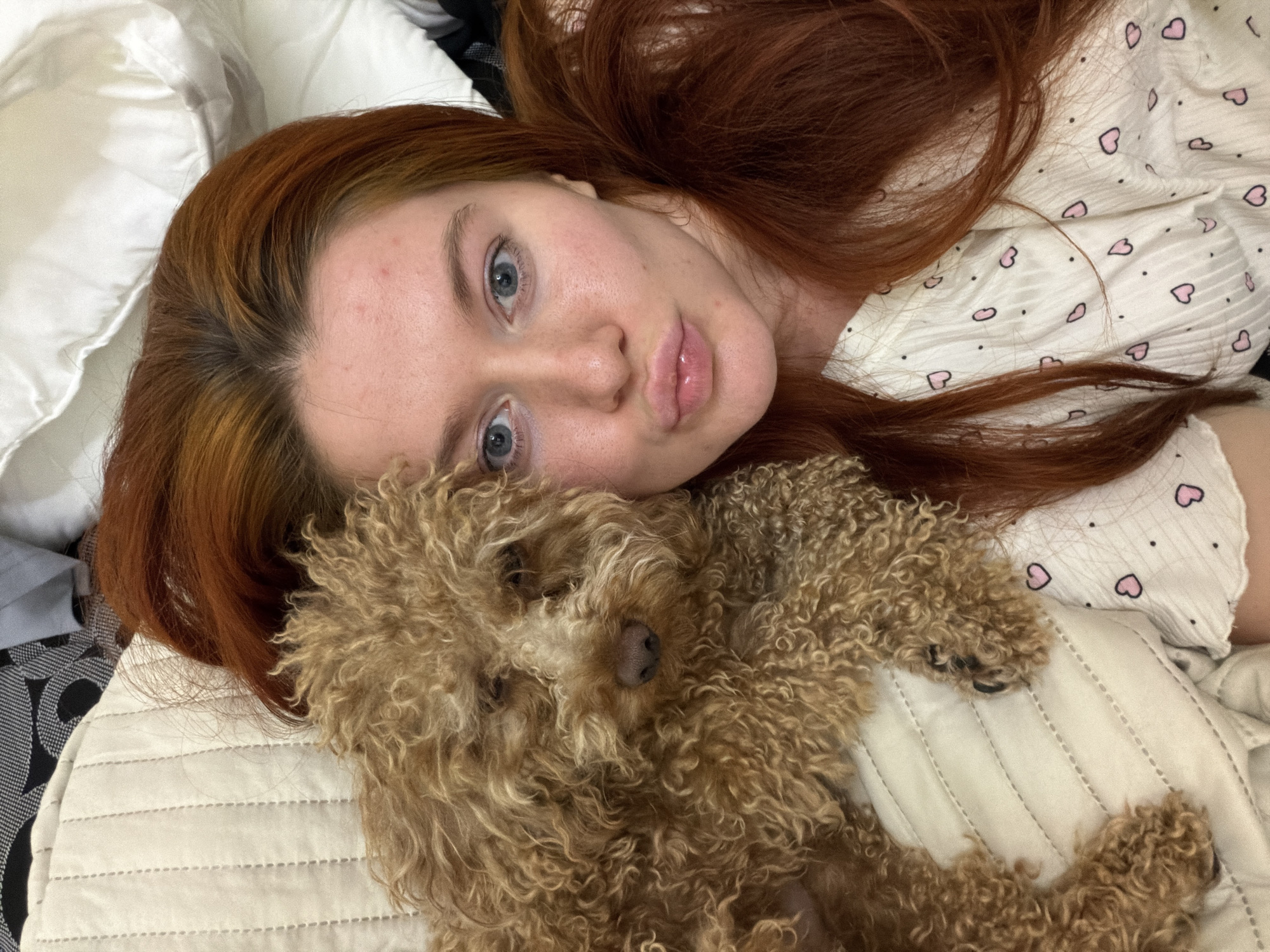 A woman with auburn hair and blue eyes is lying in bed with a small, curly-haired brown dog. The woman is wearing a white shirt with small pink hearts and black dots. She is looking at the camera with her lips puckered. The dog is resting its head on the woman's chest. The bed appears to be in a bedroom in Krasnodar, Russia.