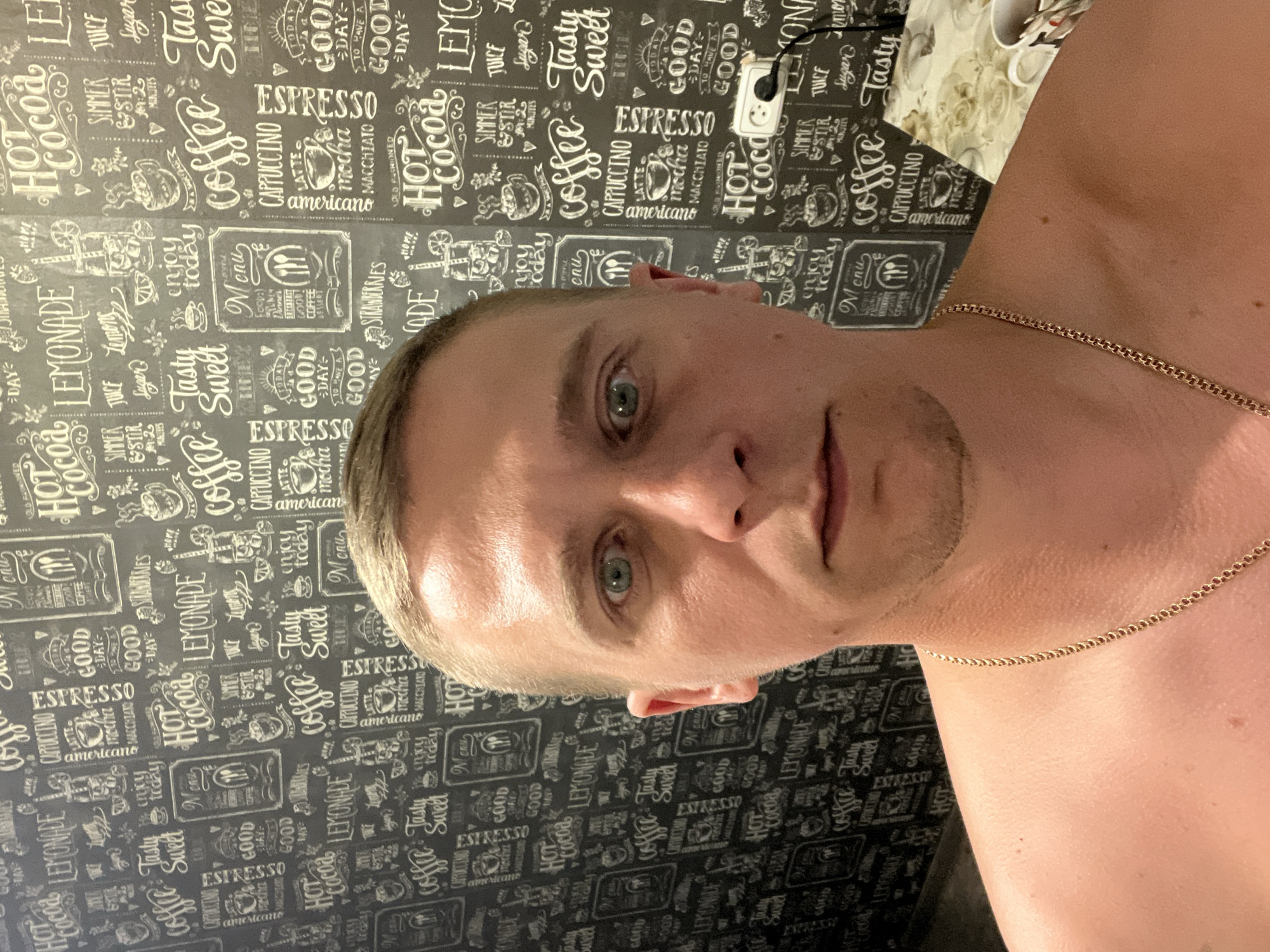 The image is a close-up, self-taken photograph of a man's face and upper chest, set against a backdrop of a patterned wallpaper. The wallpaper features a repeating design of text and illustrations related to coffee and beverages, such as "Hot Cocoa," "Lemonade," "Coffee," "Cappuccino," and "Espresso." The man, appearing to be Caucasian with light skin and short blond hair, is looking directly at the camera with a neutral expression. His right eye is a prominent light blue or green. He is wearing a gold chain around his neck. To the right of the man, partially visible, is a table with a patterned tablecloth and a mug. A white electrical outlet with a black plug is visible on the wallpaper. The lighting appears to be artificial and casts a warm glow on the man's skin. The overall impression is of a casual, indoor setting. There is no visible indication of the location being in Minsk, Belarus.