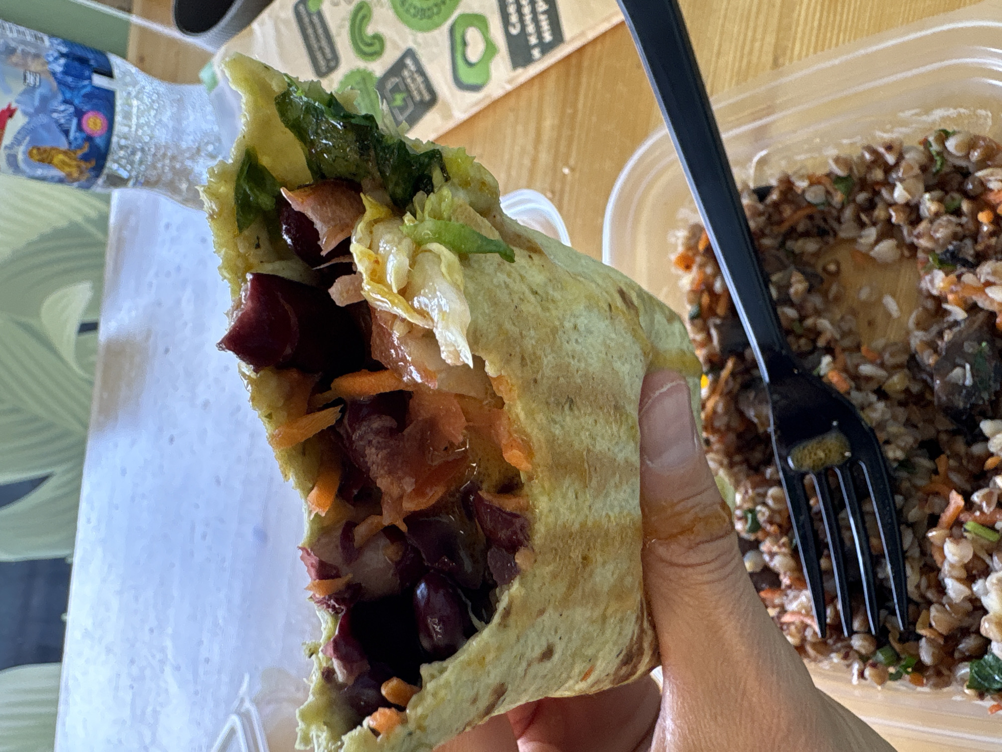 A close-up, slightly tilted image captures a person's hand holding a large, open-faced wrap filled with a colorful medley of vegetables. The wrap, appearing to be a pale green or yellowish tortilla/lavash, showcases its moist interior. Visible ingredients include dark red components, possibly kidney beans or beetroot, along with shredded orange carrots, and both light and dark green leafy vegetables such as lettuce.
To the right, a clear plastic container holds a grain-based dish, likely buckwheat, mixed with finely chopped orange carrots and specks of green herbs. A black plastic fork is resting inside this container. A plastic water bottle with a blue and white label is partially visible in the upper left background.
The meal is set on a light-colored surface. In the background, a patterned paper bag or placemat features several green, organic-shaped logos. Visible Cyrillic text on this paper item includes the words "Свеж" (meaning "Fresh") and "качество" (meaning "quality"). The bright, diffused lighting suggests an indoor setting. The image depicts a casual meal, with the person actively holding the wrap, implying they are either about to take a bite or have just done so.