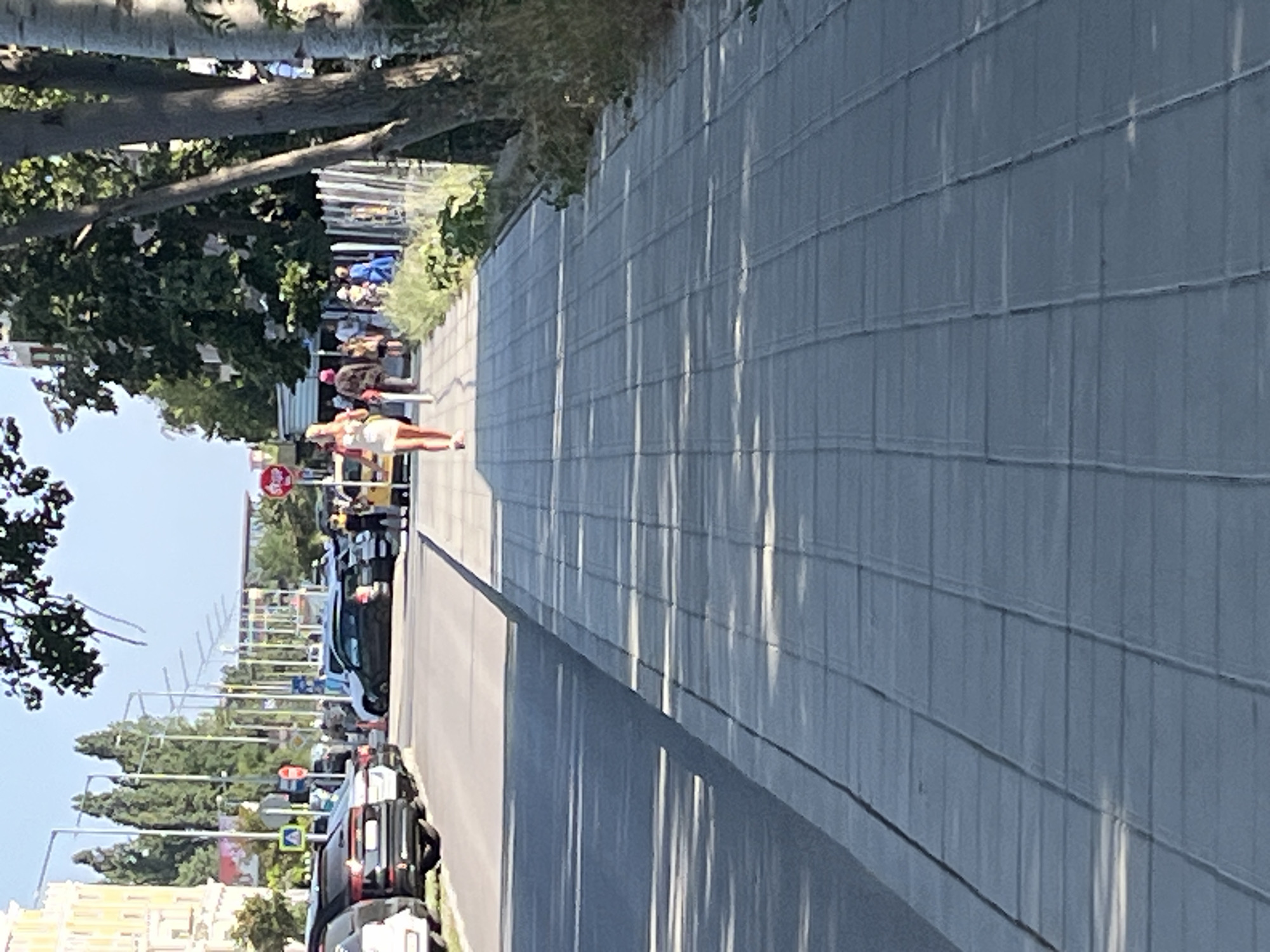 A woman in a light-colored dress and pink shoes is walking on a paved sidewalk alongside a street in Nesebar, Bulgaria. The street is lined with parked cars, including a black SUV, a silver car, and a yellow taxi. Buildings and trees can be seen in the background. The sidewalk is made of gray rectangular pavers, and shadows from trees are cast across it. The scene appears to be a sunny day.