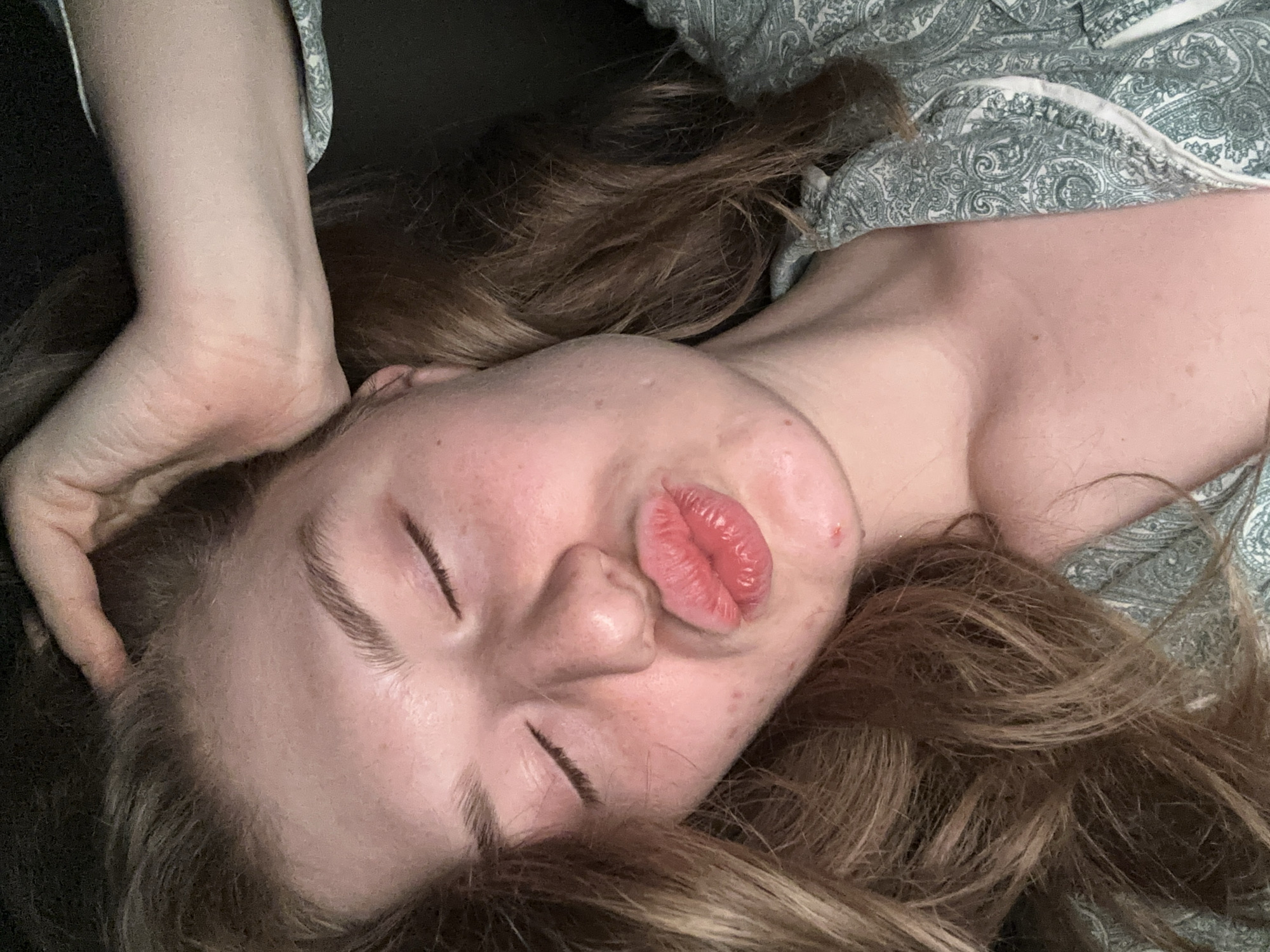 A close-up, angled shot depicts a person lying down. The individual has fair skin, closed eyes, and puckered lips. One hand rests on their head. Loose, light brown hair surrounds the face. The person is wearing pajamas with a light green paisley pattern. The background is dark and out of focus. The location is unknown and there is no information to indicate the location is in Kazan, Russia.