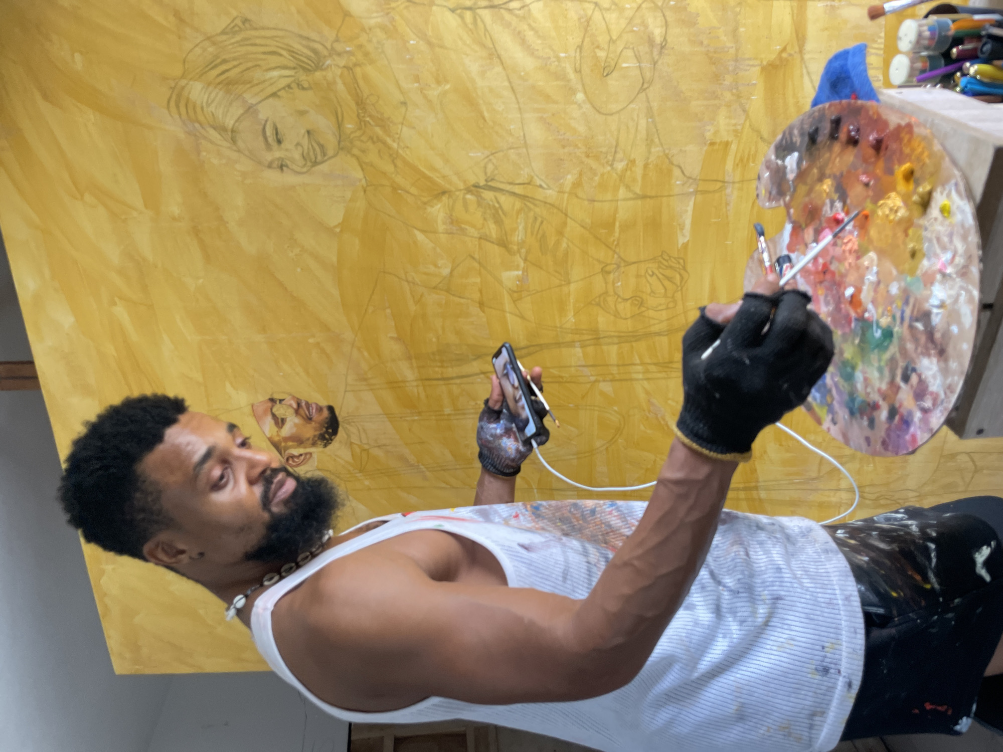 An artist in Ado, Nigeria, stands before a large, yellow canvas with a drawing of two figures. He wears a white tank top with paint splatters, black gloves, and a beaded necklace. He holds a smartphone in one hand and a paintbrush in the other. A round painter's palette, filled with various colors, is positioned to his right. Additional paint supplies are visible atop a wooden surface next to the palette.