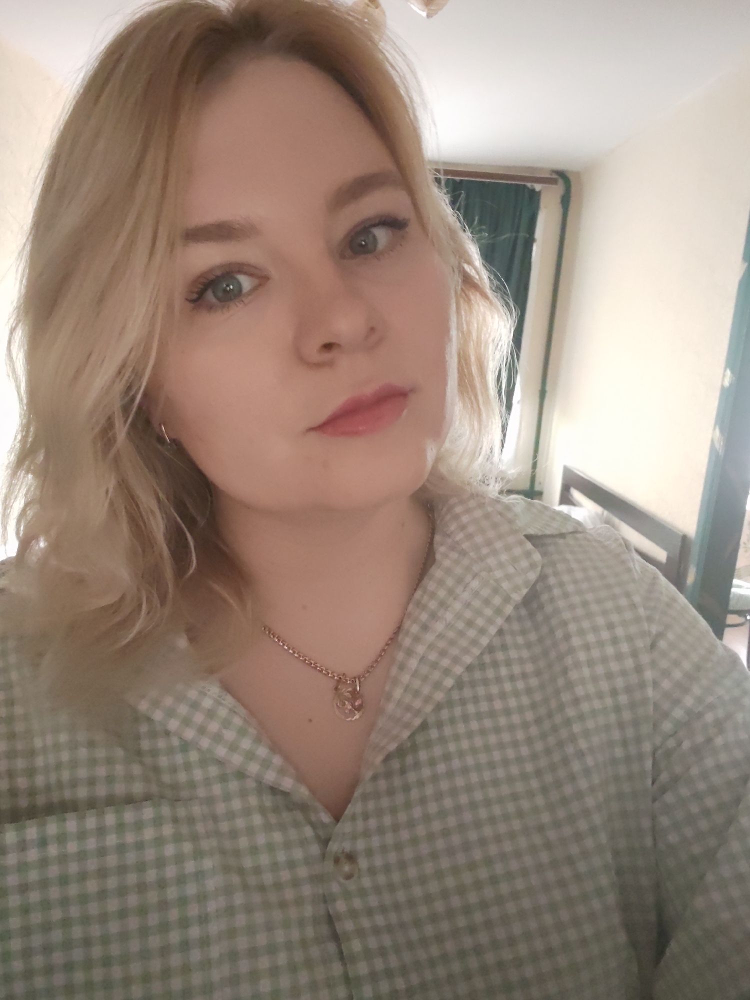 A young woman with blonde wavy hair and light skin is posing indoors, possibly in a bedroom. She is wearing a green and white gingham-patterned shirt and a gold necklace with a pendant. She is looking directly at the camera with a slightly serious expression. The background is a sparsely furnished room with beige walls, a bed frame, and green curtains partially visible through a window. There is no evidence to suggest that the location is in the city of Izhevsk, Russia.