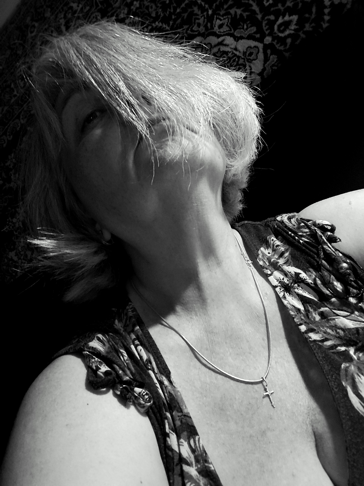 A grayscale, close-up, low-angle shot captures an overhead view of a woman looking upwards, her face partially obscured by her short, layered, blonde hair. Her eyes are visible, and her mouth is slightly open, forming an "O" shape. She is wearing a floral-patterned top and a silver chain with a small cross pendant around her neck. The background features a dark, textured pattern. The scene is dimly lit, with harsh lighting creating strong shadows. The woman's head is tilted back, and her neck is extended, revealing her collarbone and part of her décolletage. The image is a self-portrait, as indicated by the camera angle. There is no identifiable location within the city of Krasnodar, Russia.