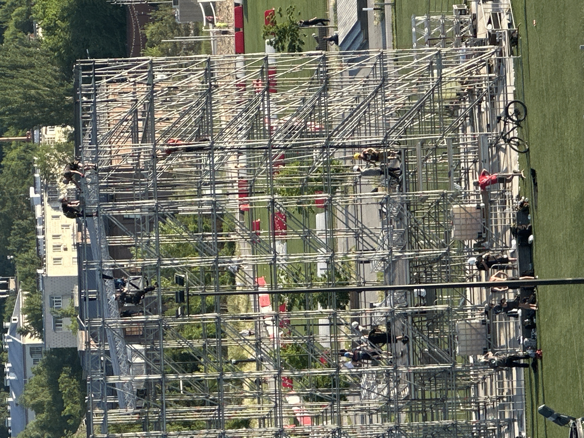The image shows an outdoor scaffolding structure in Kazan, Russia. A group of people are assembled on the ground near the scaffolding. A bicycle rests against the scaffolding. Red and white objects are interspersed within the scaffolding. The background includes trees, buildings, and a green grassy area.