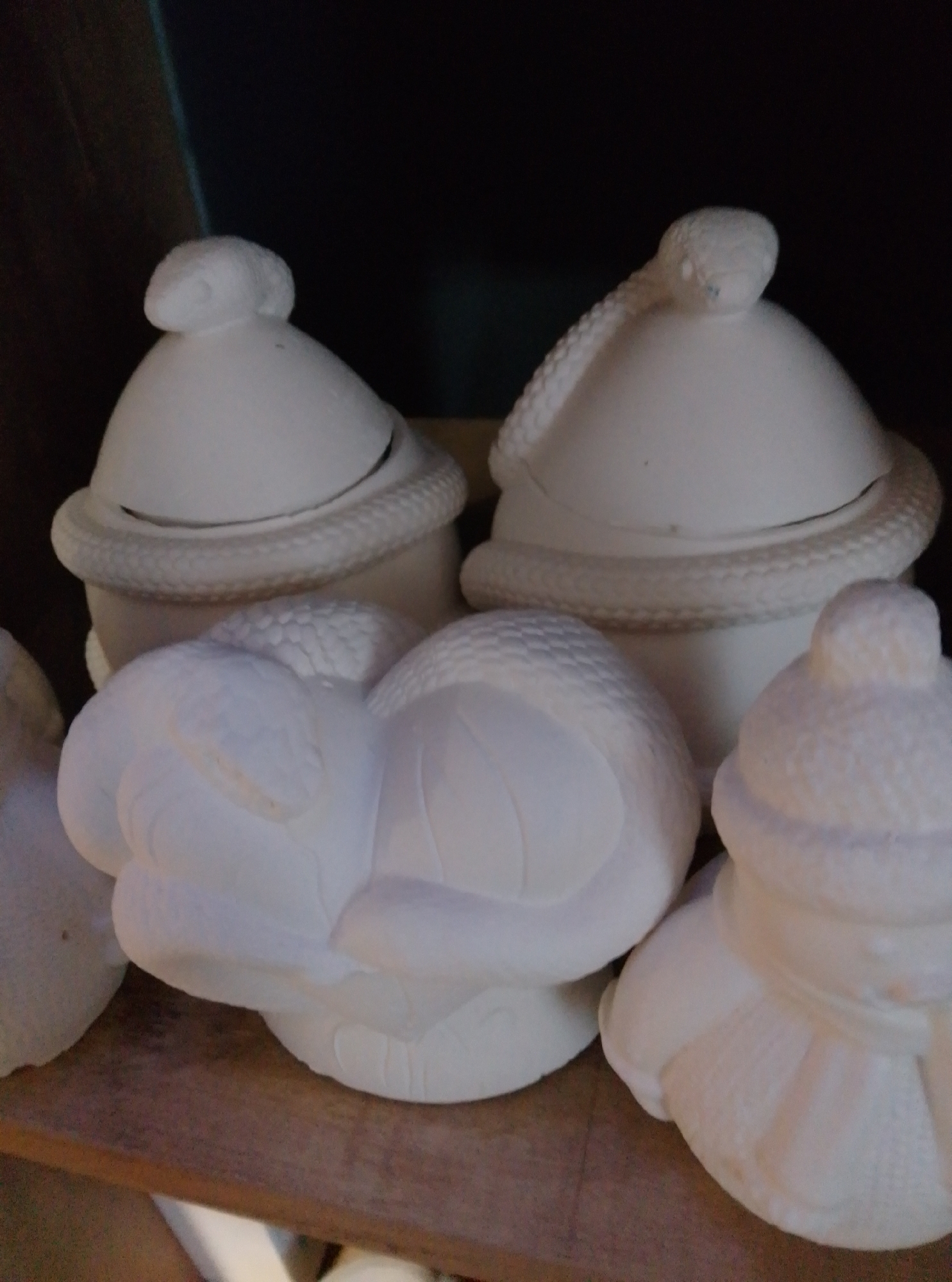 The image shows several unpainted ceramic pieces on a wooden shelf against a dark background. The objects appear to be decorative items, possibly for crafts or seasonal displays. On the left, a lidded jar with a snake coiled around its rim and head on top is visible. Next to it is another similar lidded jar, also with a snake design. In front of these jars are two other ceramic pieces: one resembles a sleeping animal, possibly a snake or a cat, curled up, and another, on the right, looks like a snowman or a similarly shaped figure wearing a hat. The ceramic pieces are white and appear to be unfinished. The image does not provide any information about the city of Wedel, Germany.