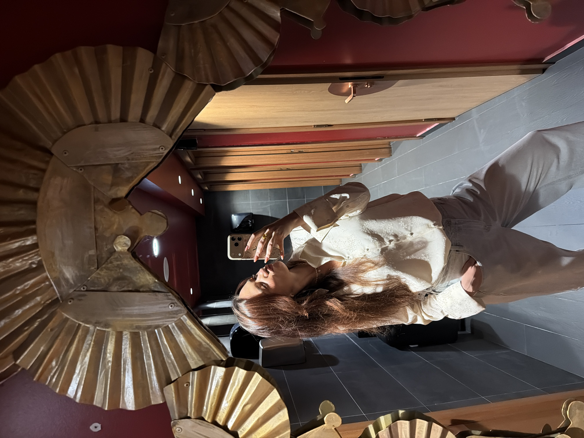 A woman is taking a selfie in front of an ornate, gold-colored mirror. The mirror appears to be in a public space, possibly a restaurant or bar in Belgrade, Serbia. The background shows a red wall and gray tile flooring. The woman is wearing a white shirt and light-colored jeans. She is holding a phone in front of her face to capture the image.
