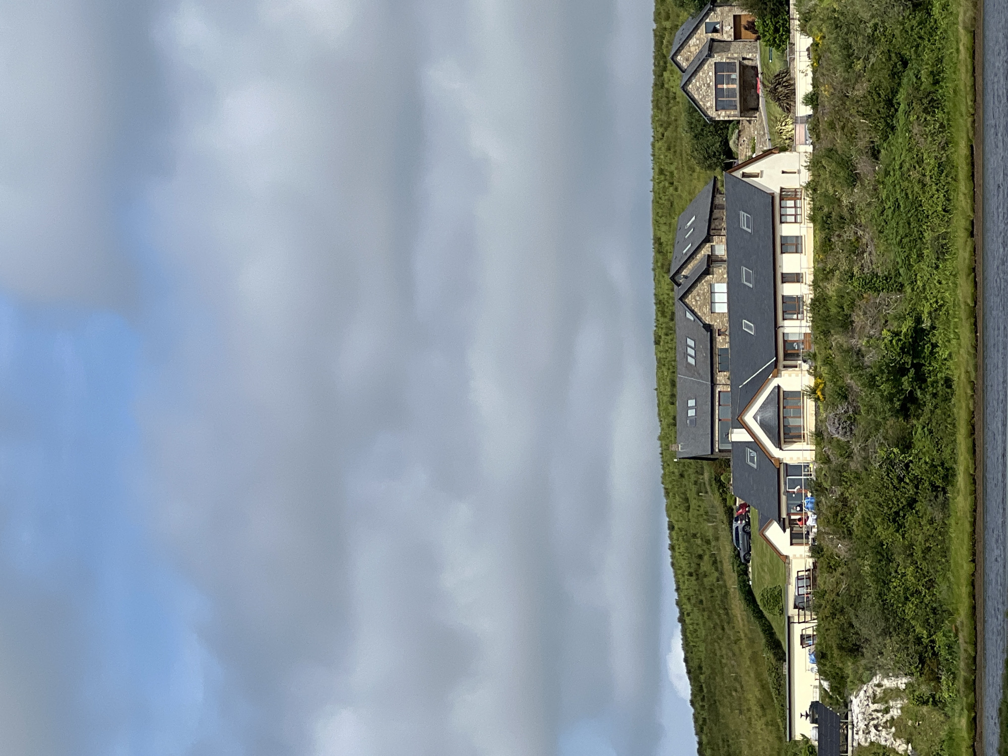 The image shows a group of modern houses situated on a grassy hillside overlooking a body of water. The architecture of the houses features stone facades and dark tiled roofs. Some houses have large windows and balconies. The surrounding landscape is characterized by rolling green hills covered in dense vegetation, with patches of rough terrain. The sky above is partly cloudy with a mix of blue sky and grey clouds. A few cars are parked on the upper part of the hillside. The foreground shows the edge of the water, with a strip of dark soil or vegetation along the bank. The scene appears to be a residential area in a rural or coastal setting.