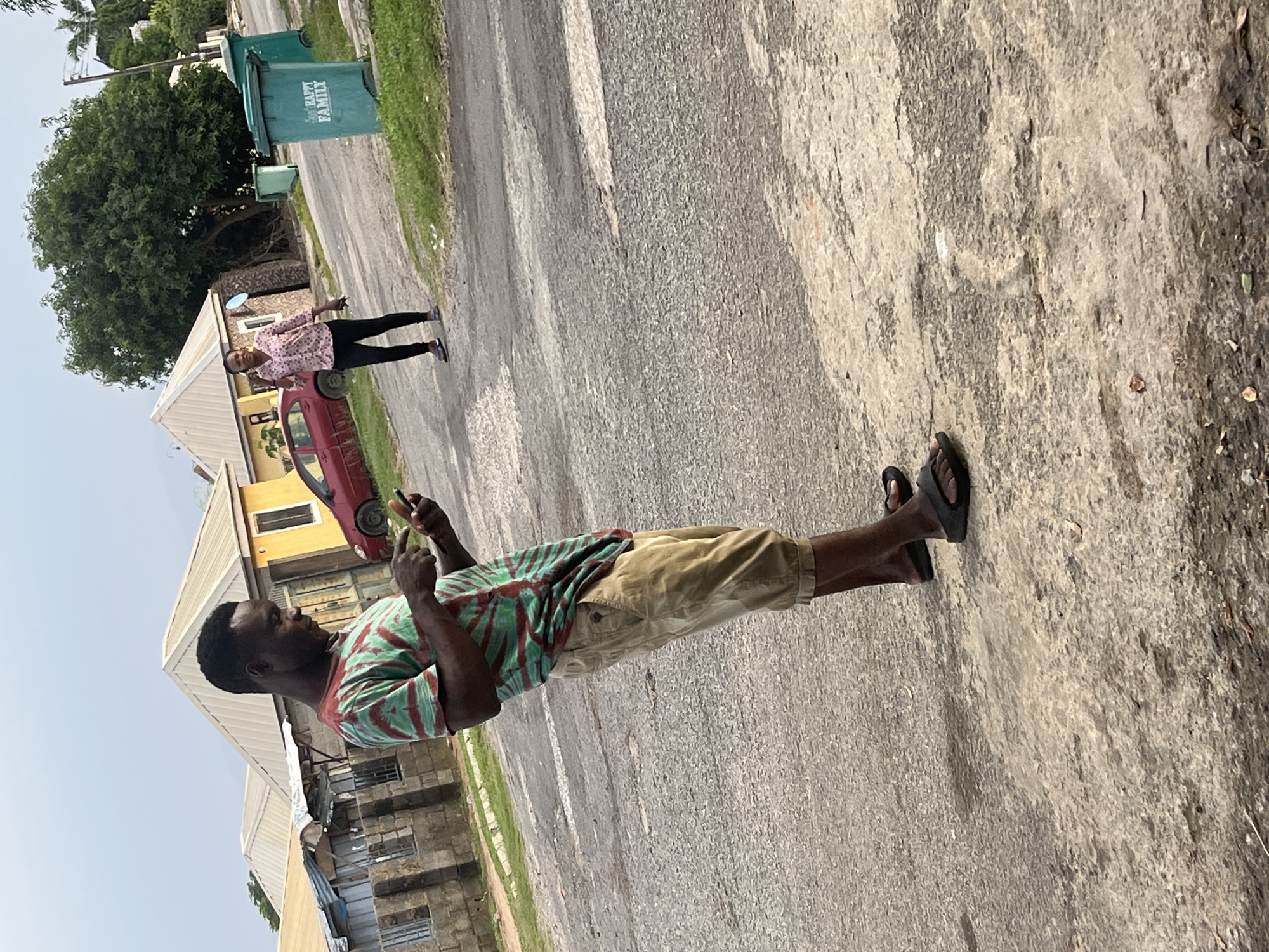 The image shows two people on a paved street in a residential area of Abuja, Nigeria. In the foreground, a man wearing a green and red tie-dye t-shirt and khaki shorts stands looking at his mobile phone. He is wearing black flip-flops. In the background, a woman wearing a pink floral shirt and black pants is walking away from the camera, also appearing to be looking at her phone. A red car is parked on the street to the left of the woman, and two green trash bins are visible on the right side of the street. The area appears to be an uncrowded residential neighborhood with houses and trees visible. The sky is overcast.