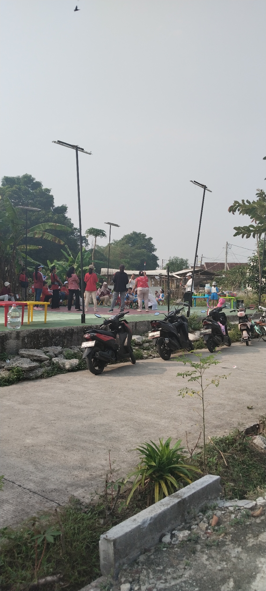 The image depicts an outdoor gathering of people in what appears to be a community space. In the foreground, two motorcycles are parked on a paved surface. Behind them, a group of people, some standing and some seated, are engaged in an activity. Several are wearing red shirts and appear to be exercising or participating in a class. One person is standing with their arms raised, seemingly leading the activity. In the background, there are trees, some buildings, and lampposts with solar panels, suggesting a developed area, possibly a park or community center. The sky is overcast. The overall scene suggests a daytime event in a semi-urban or suburban environment. The provided context indicates the location is the city of Tabanan, Indonesia.