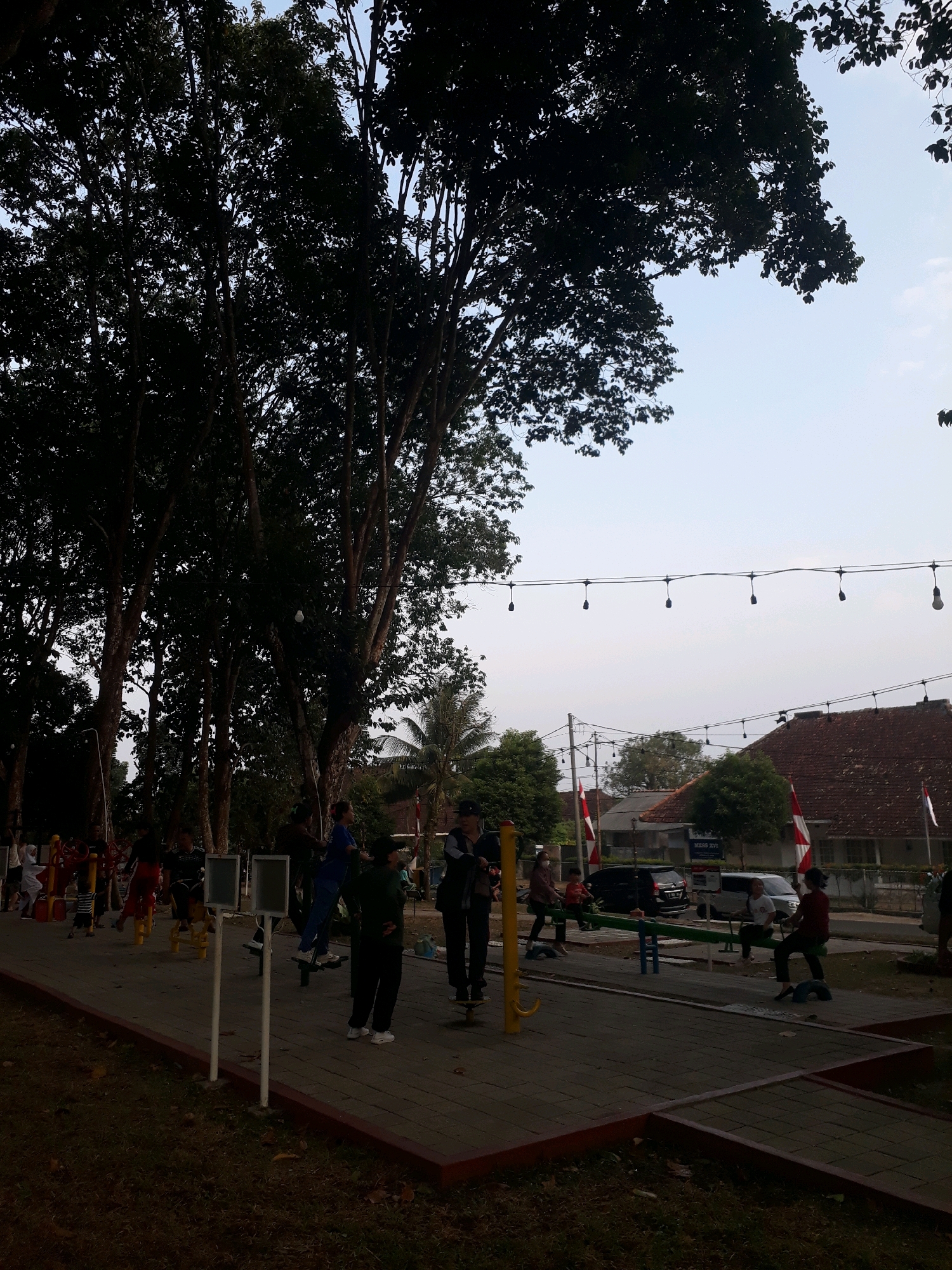 The image depicts an outdoor public park area, likely in Pangkalpinang, Indonesia, featuring several individuals engaged in physical activity. Tall trees with dense foliage dominate the left side of the frame, casting shadows. In the foreground, a paved walkway is bordered by grass. A variety of outdoor exercise equipment, including a leg press and a stationary bike, are visible. Several people are using these machines. Further into the background, there are houses with tiled roofs and Indonesian flags displayed. A string of decorative lights hangs across the sky above the park area. The lighting suggests it is either early morning or late afternoon.