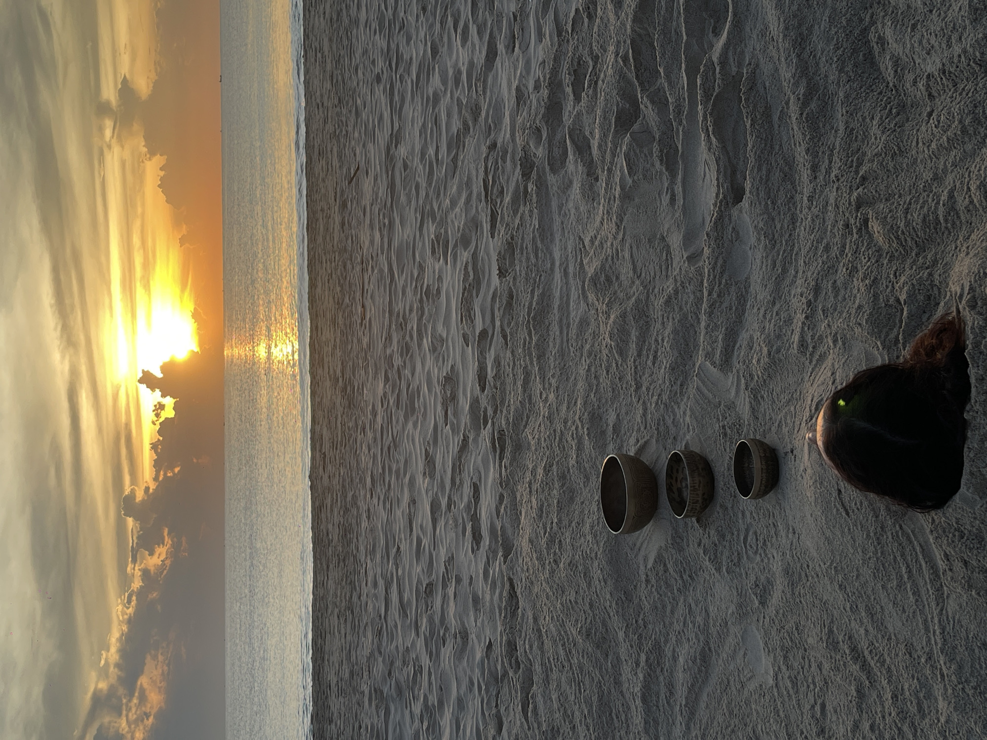 This image depicts a serene beach scene at sunrise in Đà Nẵng, Vietnam. The foreground is dominated by textured sand with footprints and three metal bowls placed in a line, partially embedded in the sand. In the bottom right corner, the back of a person's head is visible, with dark hair and a faint green glow emanating from what appears to be an eye. The middle ground features a calm sea with a reflective surface, meeting a sandy shoreline. The background showcases a vibrant sunrise with the sun partially obscured by dramatic, illuminated clouds in shades of orange and yellow. The sky transitions to a softer gradient of yellow and grey. A few distant boats are visible on the horizon, suggesting the presence of maritime activity.