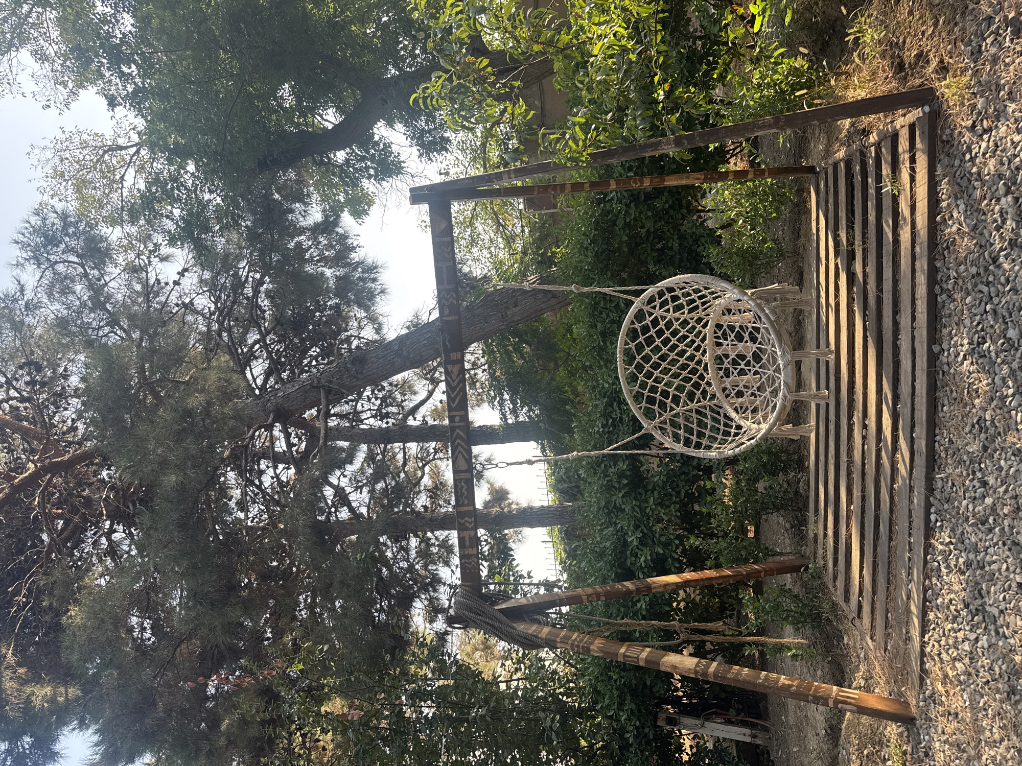 A white macrame hanging chair is suspended from a wooden swing set with a decorative beam. The swing set is situated on a wooden platform surrounded by gravel and green foliage. Tall trees with green leaves and pine needles are visible in the background under a bright sky. The scene is outdoors, suggesting a park or garden setting. The image does not depict any people or specific landmarks that would identify its location in Baku, Azerbaijan.