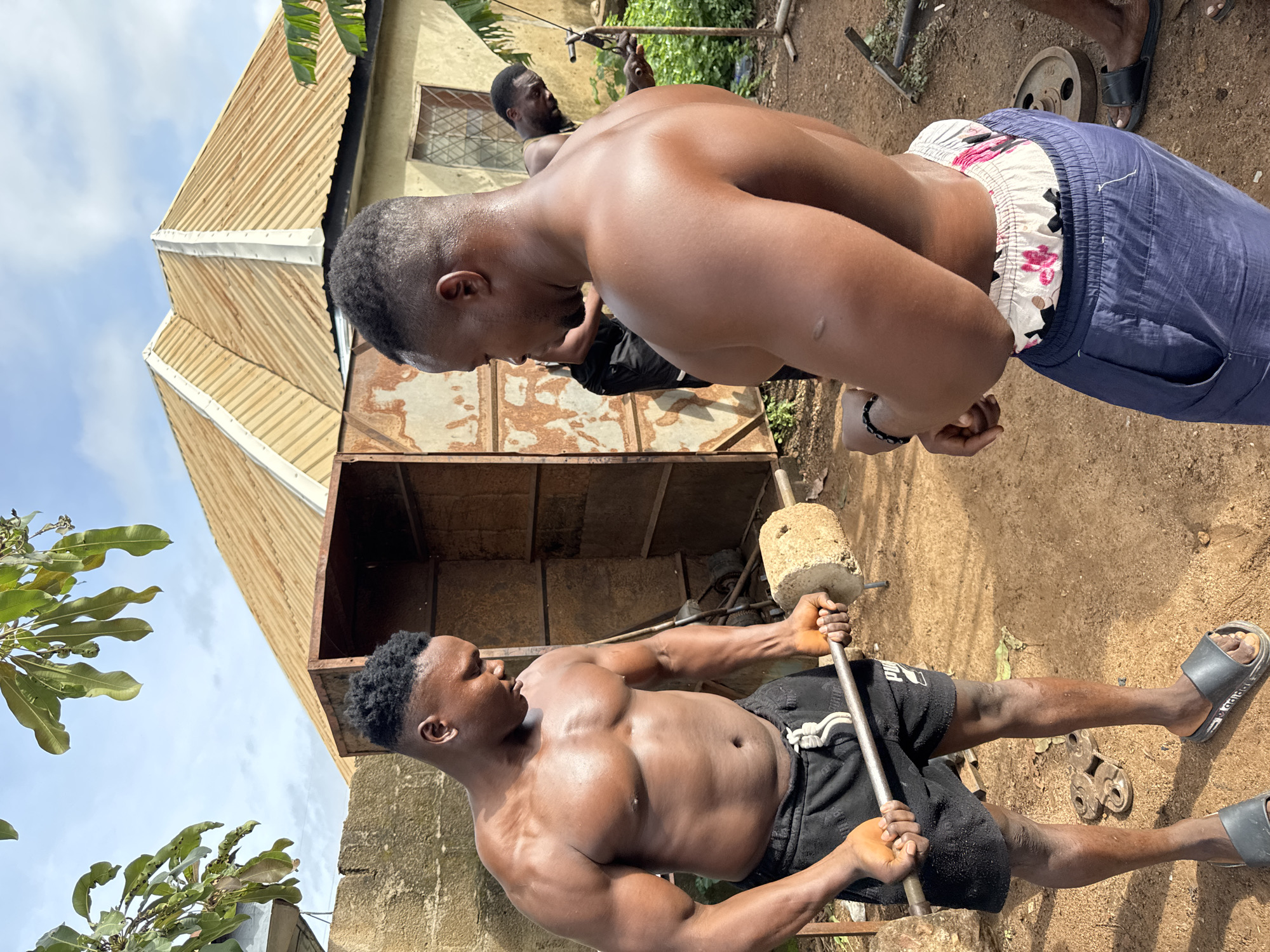 This image captures an outdoor, informal gym scene in Akwanga, Nigeria, featuring three Black men engaged in weightlifting activities under a bright, partly cloudy sky.
In the foreground on the left, a muscular, shirtless man with short, dark, textured hair stands, facing right and slightly downward. He wears black shorts with a white drawstring and black slides. He holds a homemade barbell, constructed from a metal bar with two rough, cylindrical concrete or stone discs acting as weights. Sweat is visible on his chest and arms, indicating physical exertion.
To the right, another muscular, shirtless man stands, oriented with his back partially to the camera, looking towards the left foreground. He has short, dark, textured hair and wears dark blue athletic pants with a floral-patterned boxer short waistband visible above them. A black beaded bracelet is on his left wrist.
In the background on the far right, a third Black man with a beard is partially visible, wearing a dark top, operating what appears to be a makeshift cable or pulley exercise machine fashioned from metal components.
The ground is reddish-brown dirt, with various discarded metal objects, including a circular metal weight plate, scattered across it. In the mid-ground, behind the left man, a rusty, open-sided metal structure, possibly a storage unit or shed, is present. Behind the men, a light-colored building with a multi-pitched, corrugated metal roof (brownish-yellow) and a window secured with metal grilles is visible. Green foliage, including large banana-like leaves, is present on the right, and tree leaves overhang from the top left.