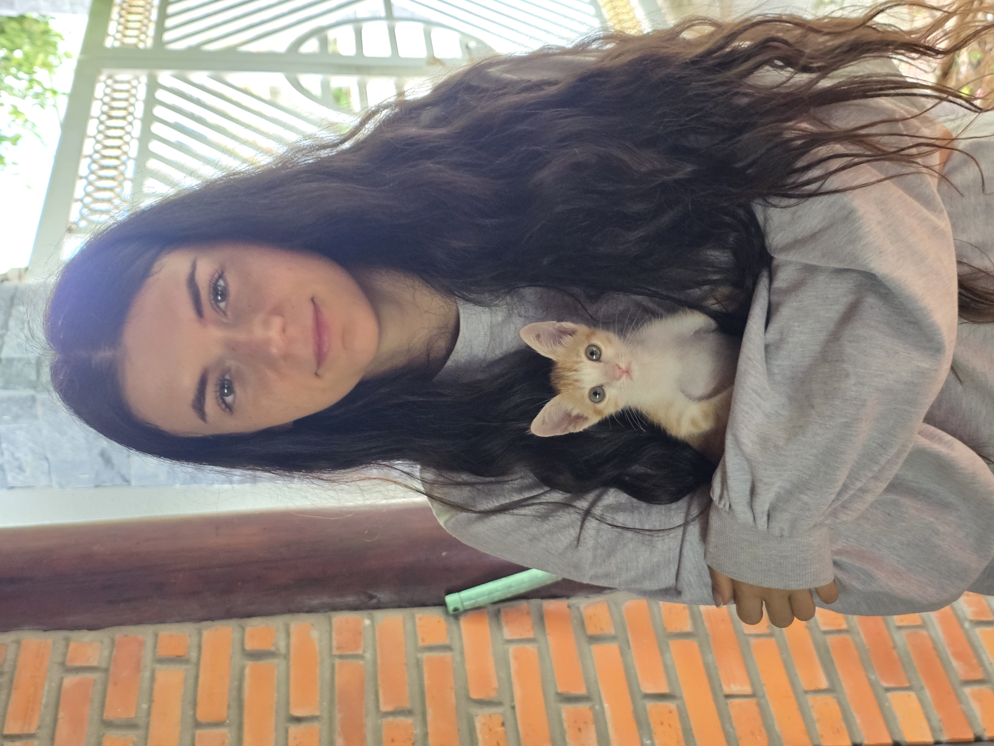 The image shows a young woman holding a small kitten, captured in what appears to be an outdoor or semi-outdoor setting in Mũi Né, Vietnam. The woman, with long, wavy dark brown hair cascading over her shoulders, looks directly at the viewer with a calm, gentle expression, and a subtle smile. She has light-colored eyes and is wearing a light grey, long-sleeved top or sweatshirt.
Cradled in her arms is a small, adorable ginger and white kitten. The kitten has distinct orange tabby markings on its head and back, with a white belly and paws. Its eyes are a striking green, and it is looking slightly away from the camera.
The background provides contextual clues about the location. Above the woman's head, a white, ornate lattice structure is visible, suggesting a decorative railing or a part of an open-air building like a gazebo or veranda. Below her arm, a dark wooden beam runs horizontally across the mid-ground. The lower portion of the image features a wall constructed from orange-red bricks, laid in a staggered pattern, adding to a rustic or traditional aesthetic. A small, light green object is partially visible beneath the wooden beam.
The scene is lit by natural daylight, indicating it is daytime, and the overall mood is serene and affectionate, highlighting the bond between the woman and the kitten. There is no visible text in the image.