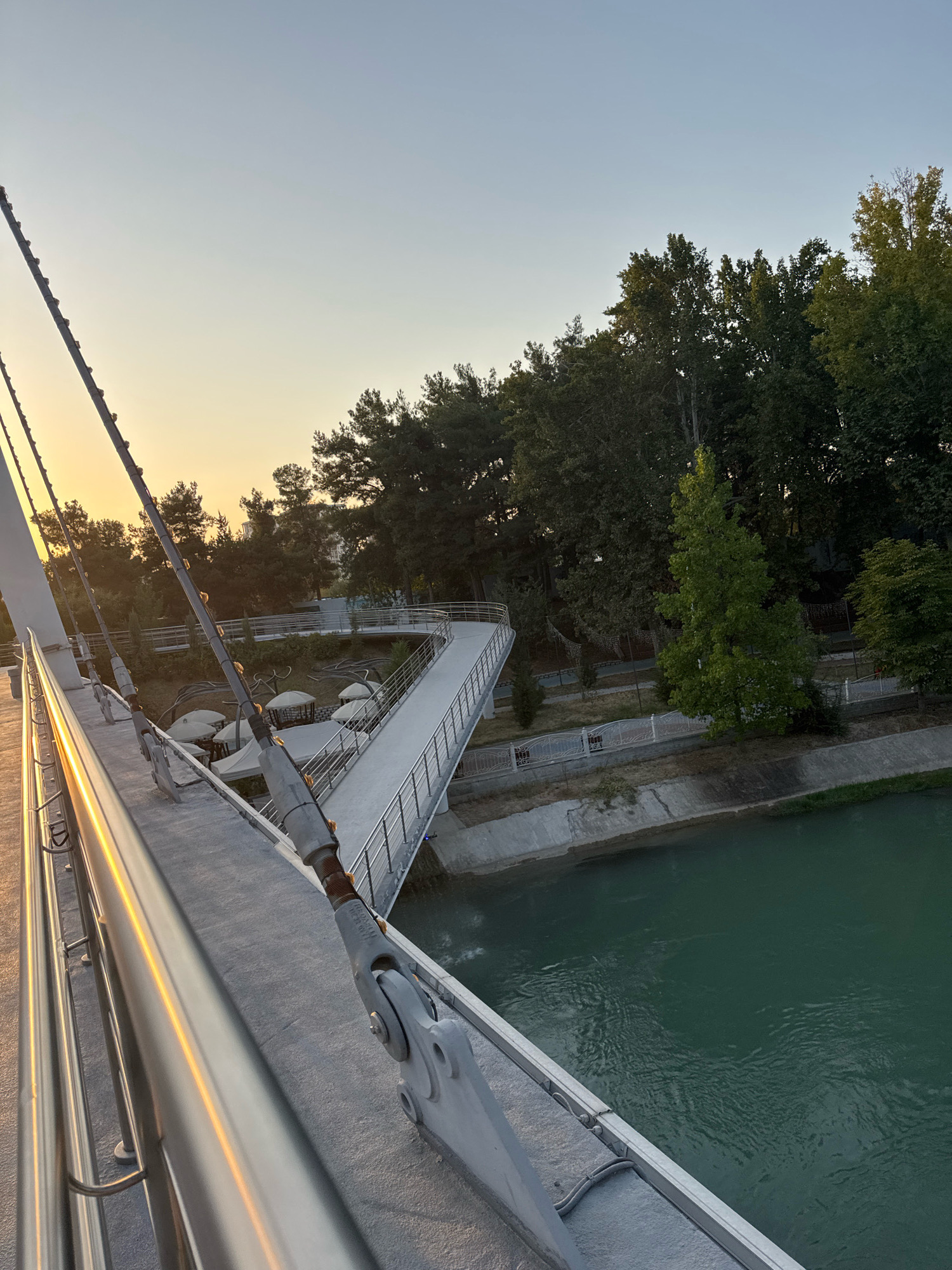 The image shows a modern pedestrian bridge in Tashkent, Uzbekistan, likely near the Tashkent TV Tower or the Monument of Independence due to the presence of mature trees and a canal. The perspective is from the bridge's walkway, looking downwards and across at a curve in the bridge and the surrounding parkland. The sky is clear with a pale yellow sunset glow on the left horizon.
The bridge itself is constructed from white concrete and metal. A silver railing runs along the walkway, reflecting the golden light of the setting sun. Several thick, steel cables, which support the bridge, are visible in the foreground, attached to large metal anchor points.
Below the bridge, a green canal flows. On the opposite bank, a well-maintained park area is visible. It features a winding pathway, shaded seating areas with white canopies, and a variety of trees, including mature pines and deciduous trees. A white metal fence separates the park from the canal bank, which is lined with concrete. No people are visible in the frame. The overall impression is one of a tranquil urban park setting at dusk.