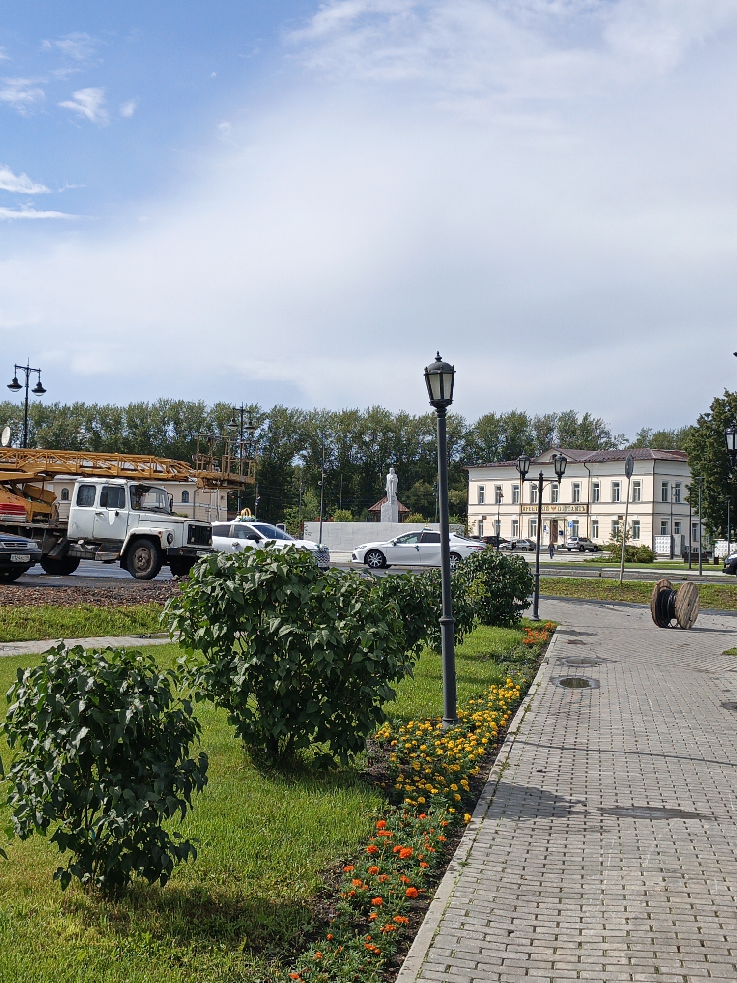 The image depicts a street scene in Irbit, Russia, with a focus on a sidewalk lined with bushes and flowers. In the background, a light-colored, two-story building with numerous windows and a sign that reads "ПРЕЗИДЕНТСКИЙ ПОЧТАМТ" (Presidential Post Office) is visible. A statue of a person stands to the left of the building. A white crane truck is parked on the road in front of the building, with several cars also present. The sidewalk is paved with grey bricks, and a lamppost stands on the left side of the image. To the right of the sidewalk, there is a large wooden spool. The sky is blue with scattered white clouds.