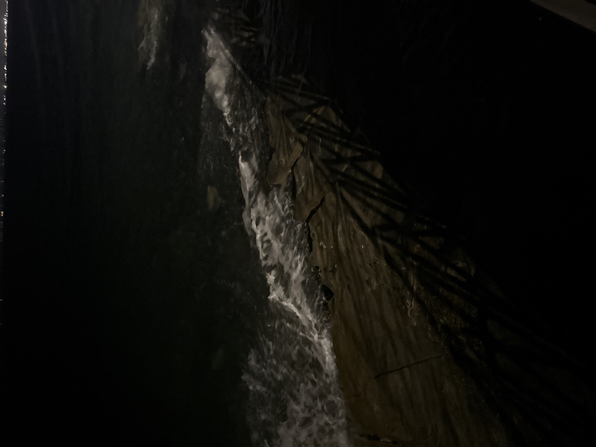 A nighttime scene captures the edge of the sea in Agios Tychona Community, Cyprus. Waves gently lap against a rocky shoreline. The water is dark, with some white foam visible where the waves break. Distant lights from a city or town twinkle on the horizon. Shadows of palm fronds are cast across the rocks and into the water, suggesting the presence of nearby vegetation. The overall impression is one of a tranquil, natural setting illuminated by artificial light sources.