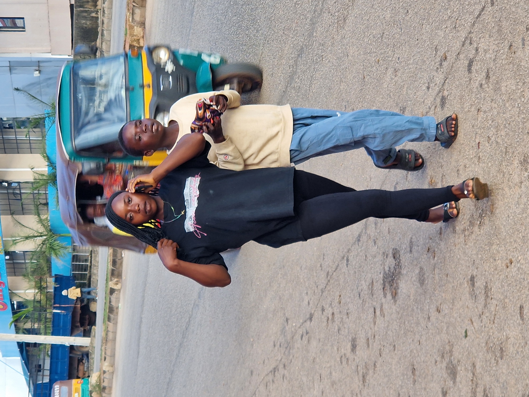 In New Stadium, Nigeria, two individuals are walking on a paved road. The person in the foreground, on the left, is a Black woman with long braided hair, wearing a black t-shirt and black leggings. She is holding a mobile phone to her ear and appears to be speaking. She is wearing black sandals. To her right, a young Black person, possibly a boy, is walking and holding a colorful object in their hands. This individual is wearing a light-colored long-sleeved shirt and light blue cargo pants, and is wearing dark sandals. In the background, a green and yellow auto-rickshaw is visible, with people inside, and a blue truck is parked further back. The environment appears to be an urban street with buildings and palm trees visible behind the road.
