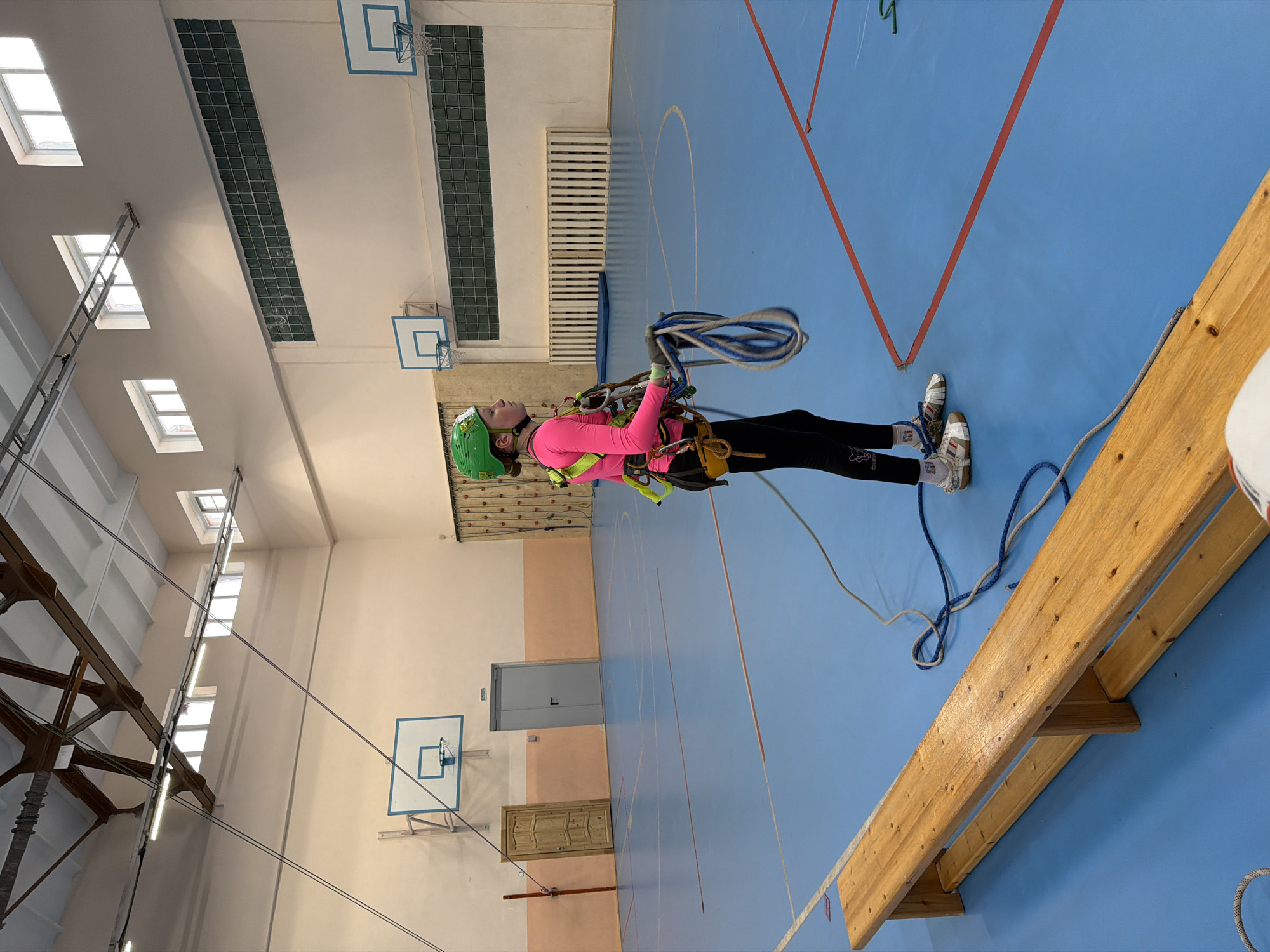 A young woman stands in a gymnasium, possibly in Berezniki, Russia, based on the gym's style and equipment commonly found in Russian schools. She is wearing a green helmet, a bright pink long-sleeved shirt, black leggings, and athletic shoes. She is also equipped with a climbing harness and ropes, holding a coiled rope in her hands.
The gymnasium has a blue floor with red lines, presumably for sports such as basketball. The walls are a light peach color. Several basketball hoops are mounted on the walls. Windows are situated along the top of the walls. A wooden bench is placed on the side.