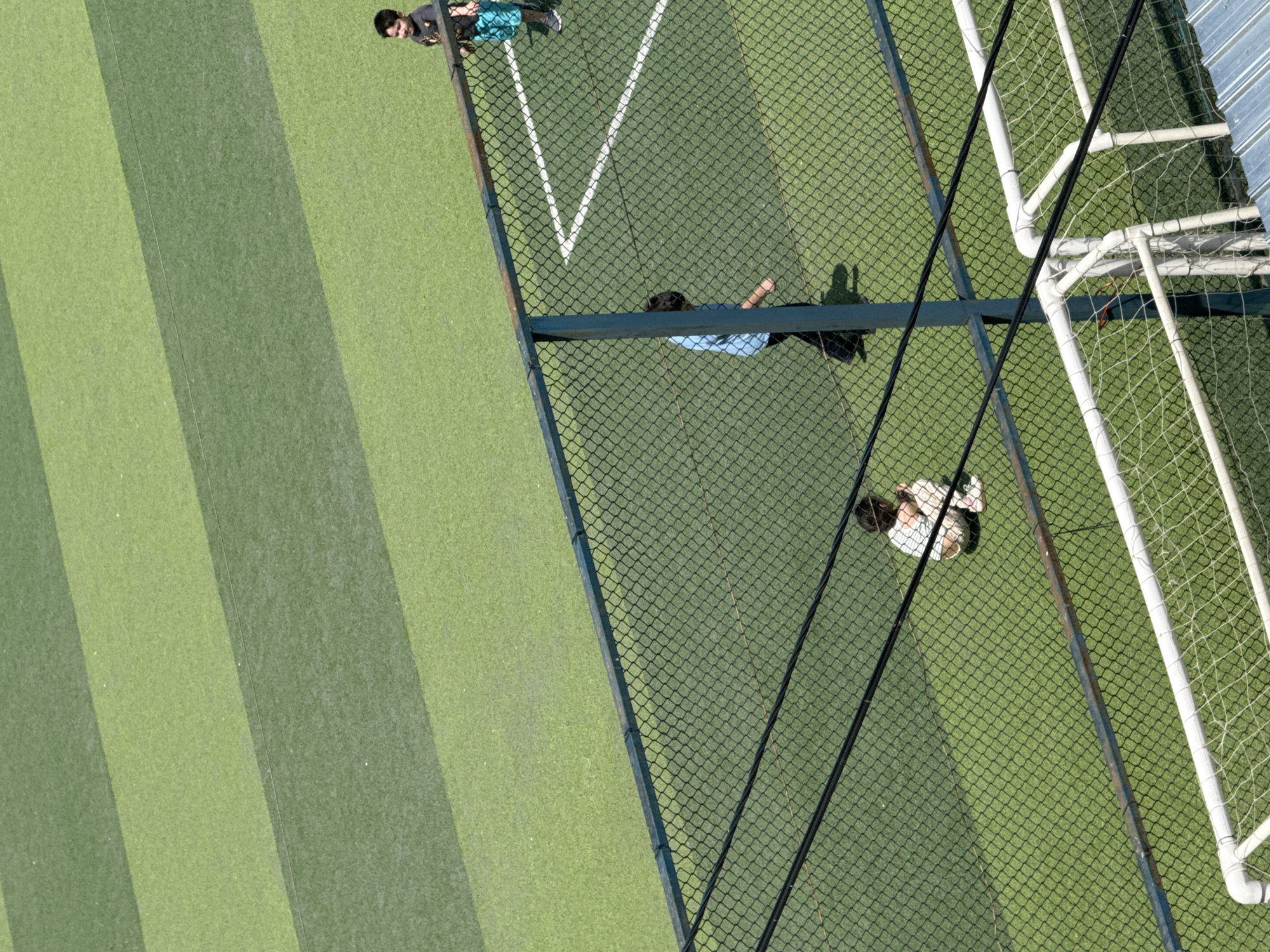 An aerial perspective shows a group of children playing on a striped green artificial turf field. In the foreground, a black chain-link fence with horizontal black wires obscures the lower portion of the scene. Three children are visible on the field. On the right side, a child wearing a dark t-shirt and blue shorts stands near a white sideline marking. In the middle of the field, another child in a light blue shirt and dark pants walks away from the camera. On the left side, closer to the fence, a child in a light-colored shirt and light-colored shorts is crouched down. A white soccer goal with a net is partially visible in the bottom right corner. The field is sectioned into alternating stripes of darker and lighter green turf, creating a geometric pattern. The environment appears to be an outdoor sports facility, likely a soccer field, under bright daylight. The location is identified as Tbilisi, Georgia.