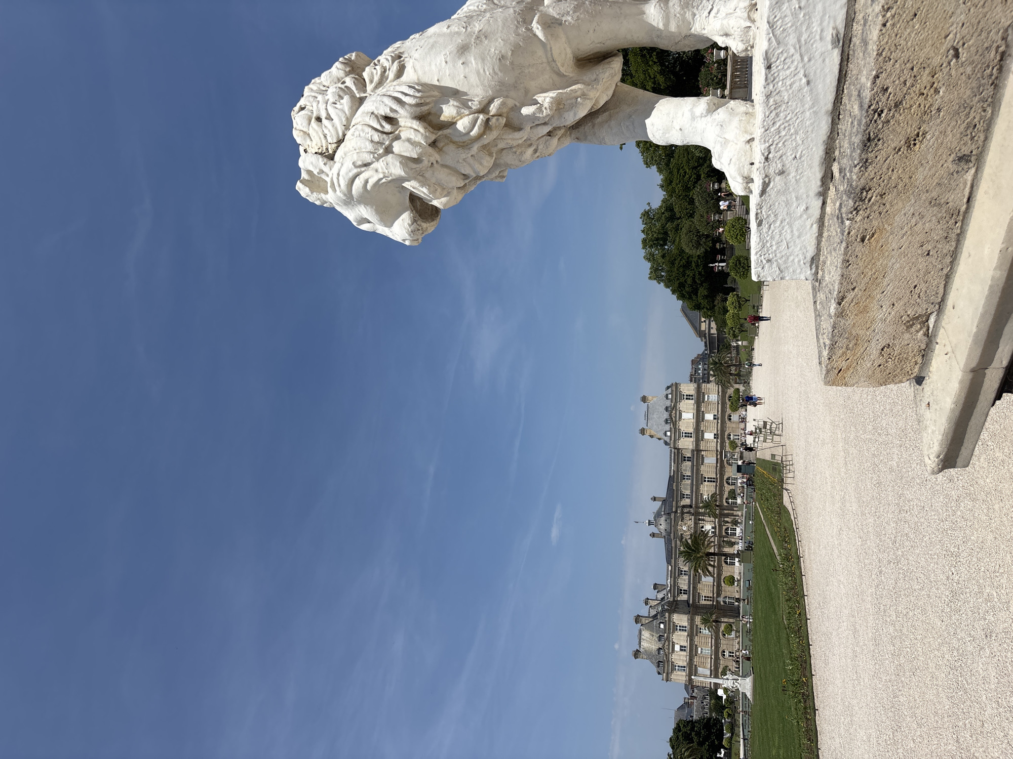 In Paris, France, a white stone lion statue stands in the foreground, its head turned to the right. Behind the statue, a large, ornate building with a symmetrical facade and mansard roofs is visible. The building is surrounded by lush green lawns, manicured trees, and a pathway made of light-colored gravel. Several people are walking along the path and in the garden area. The sky is a clear blue with a few wispy clouds. The image appears to be taken in the daytime.