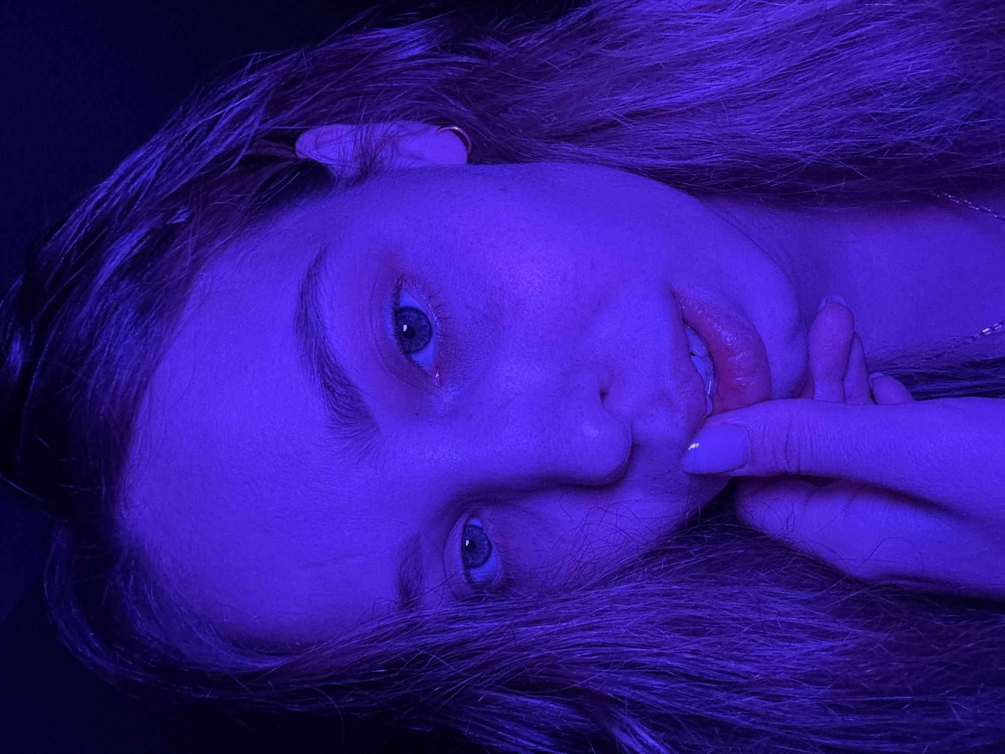 A close-up, tilted portrait photograph shows a fair-skinned person with light brown hair. The lighting is predominantly blue, casting a cool tone over the entire scene. The person is looking directly at the camera, with their right hand near their lips. The background is dark and indistinct. There is no information available to suggest this image was taken in the city of Chaikovsky, Russia.