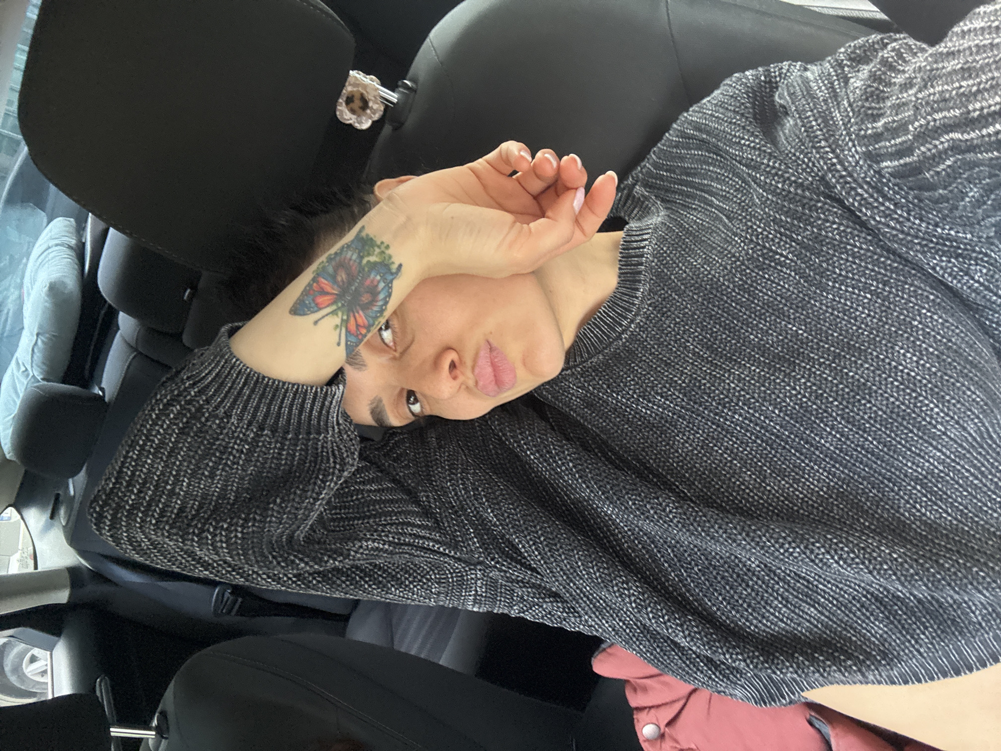 The image depicts a young woman relaxing inside a car, presumably in Tbilisi, Georgia. She is shown from the chest up, positioned on her side or leaning back, looking towards the camera with a playful, slightly pouting expression. Her right arm is bent, with her hand held near her face, possibly shielding her eyes from bright light or simply posing.
She is wearing a textured, dark grey knit sweater. A distinctive and colorful butterfly tattoo, featuring shades of red, blue, and green, is prominent on her left forearm. Her fingernails are painted a light, neutral color.
The setting is the interior of a car, characterized by black leather seats and headrests. A decorative, heart-shaped accessory, possibly with a leopard print design, is attached to the passenger-side headrest behind her. A glimpse through the car window suggests a daytime outdoor environment, though the view is blurred. A reddish-pink piece of clothing or accessory is visible in the bottom right corner of the frame. There is no visible text in the image. The overall impression is one of casual relaxation in transit.