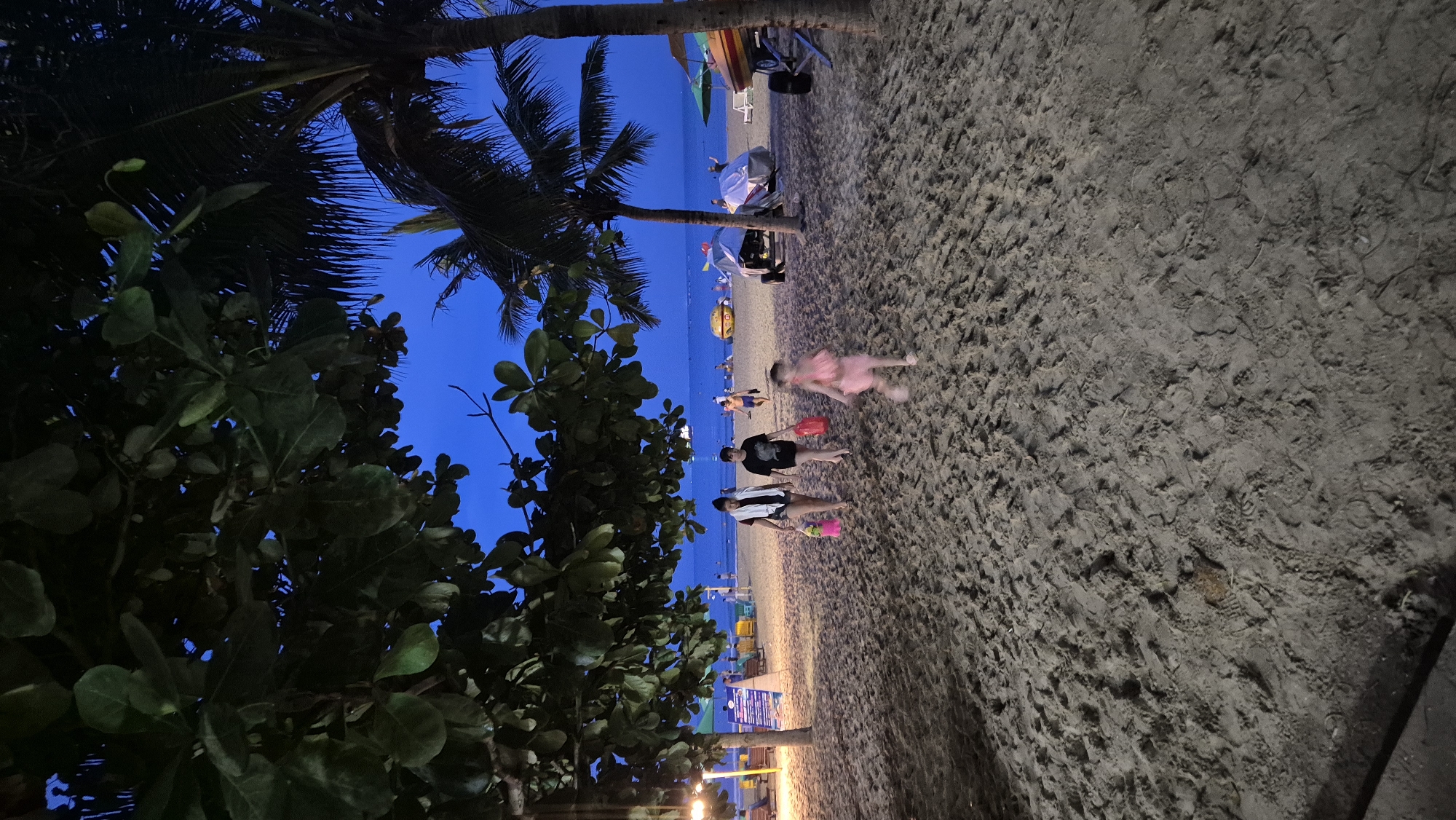 The image depicts a beach scene at dusk, likely in Đà Nẵng, Vietnam, as suggested by the presence of palm trees and a tropical beach environment. In the foreground, a sandy beach stretches out, marked with footprints. Two women are walking towards the left, carrying bags. One woman wears a white towel draped over her shoulders and carries a pink bag, while the other wears a black t-shirt and shorts and carries a red bag. A young child in a pink dress is running ahead of them towards the right. In the background, the ocean is visible under a deep blue twilight sky. Several beach chairs and umbrellas are set up, and some boats and watercraft are parked on the sand. Other beachgoers can be seen scattered in the distance. The overall atmosphere is relaxed and suggests the end of a day at the beach.