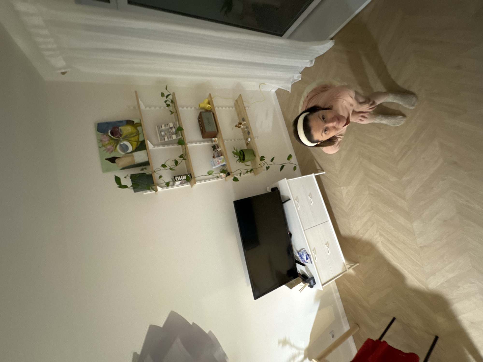 The scene is a high-angle interior shot. A person with a white headband is standing and looking up toward the camera. The floor appears to be light-colored parquet. There is a TV mounted on the wall near a white cabinet with drawers. A wooden shelving unit with plants and decorative items is on the wall. A window with white curtains is at the top of the image. A red chair can be seen at the edge of the shot. The location is not identifiable as being in Izhevsk, Russia, from this image alone.