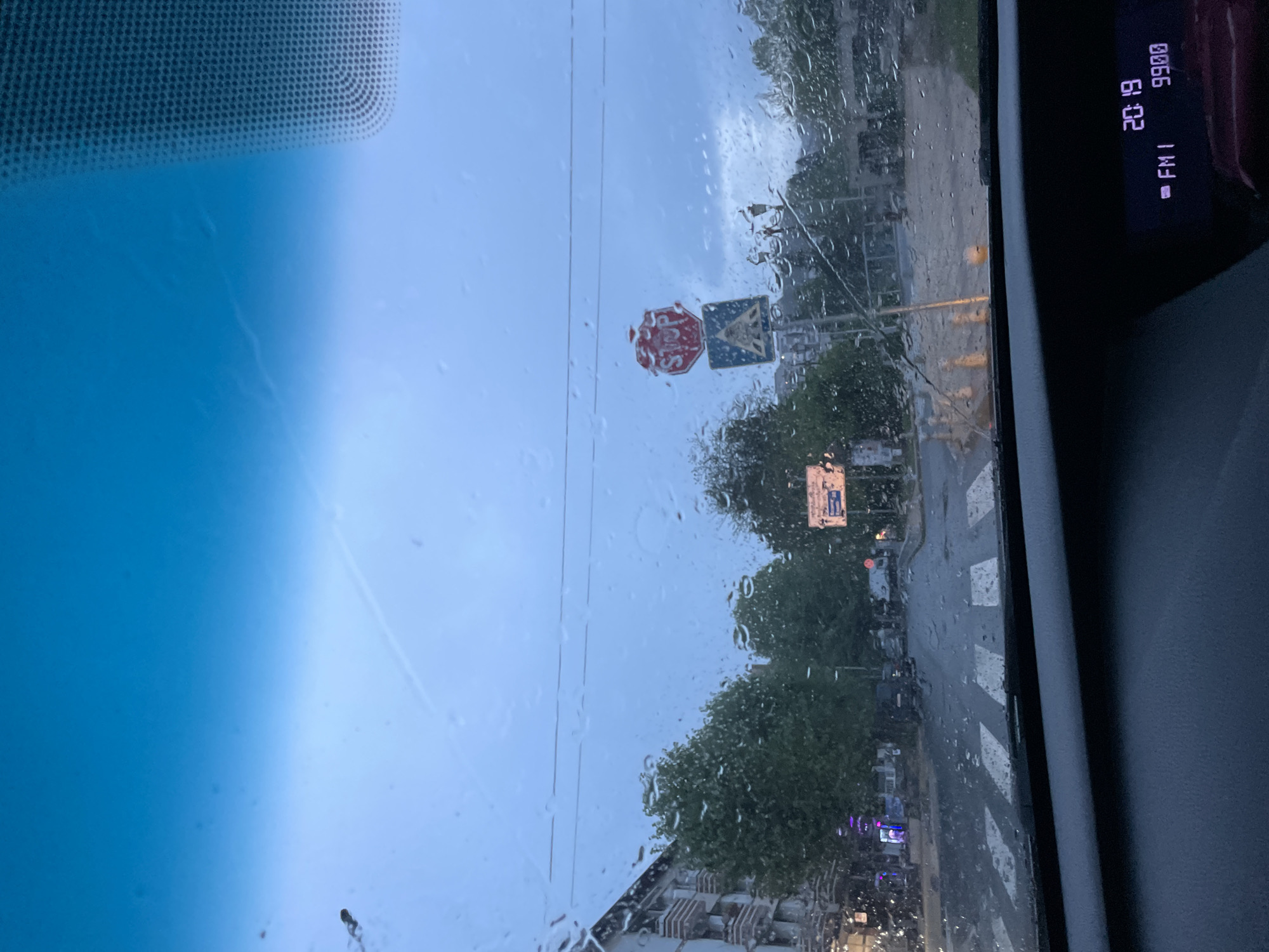 Captured from inside a vehicle, the image presents an oblique view through a rain-streaked windshield. The foreground is dominated by the vehicle's interior elements: the upper portion of the windshield exhibiting a blue gradient, and the edge of the car's console displaying digital information including "20:19" and "FM1 9900."
The focus shifts to the exterior scene, characterized by a street view under overcast skies. Raindrops obscure details but shapes are visible, particularly a "STOP" sign and an additional road sign that are adjacent to one another. The surrounding environment includes trees, buildings, and street lights, indicative of an urban setting. The image gives the impression of a street in Zlatograd, Bulgaria.
