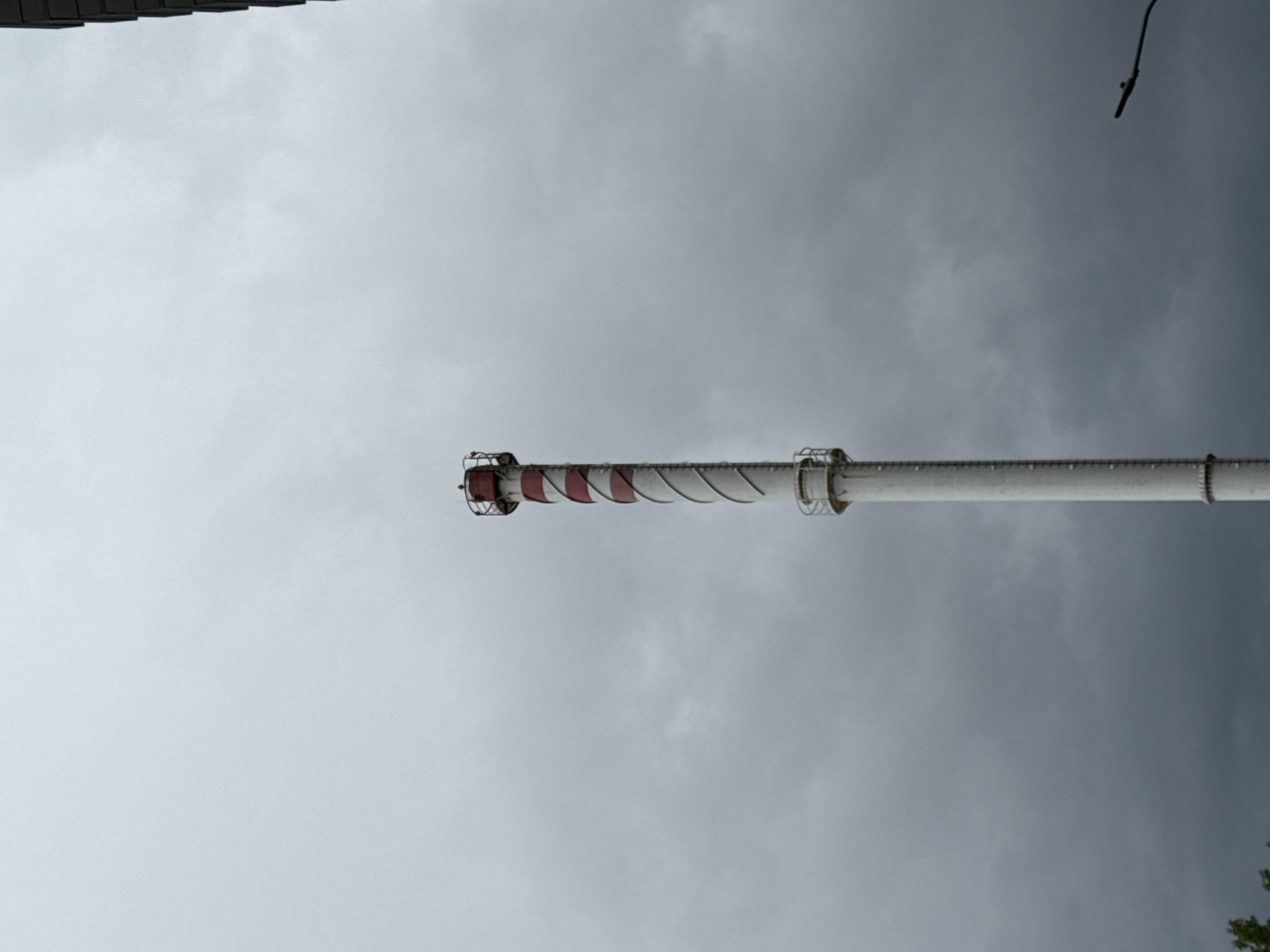 The image depicts an industrial chimney with red and white markings, set against a gray, overcast sky. The chimney appears to be part of a heating or industrial complex, given its design and location. The exact location within Moscow is difficult to ascertain from the image alone, but the presence of a chimney suggests an area with industrial or utility infrastructure. Other elements include parts of trees at the bottom right.