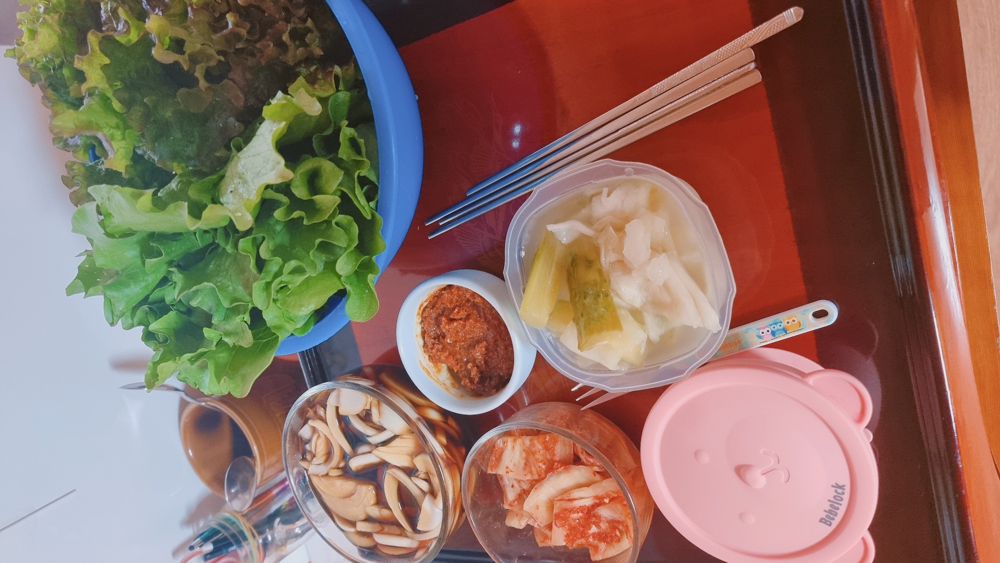 The image shows a meal laid out on a surface with a reddish-brown hue and dark stripes, possibly a wooden tray. In the upper left, there is a blue bowl containing a variety of green and reddish lettuce leaves. To the right of the bowl are two sets of chopsticks, one of a light brown color and the other of a metallic blue. There are four small containers, arranged roughly in a square:
- A small, white bowl holding a reddish-brown paste, likely a type of chili sauce or condiment.
- A plastic container filled with a mixture of white and greenish-yellow pickled vegetables.
- A glass bowl containing thinly sliced mushrooms in a dark liquid, possibly soy sauce.
- A glass bowl filled with sliced kimchi, recognizable by its bright red color.
The container in the bottom right is covered with a pink, bear-shaped lid labeled "Bebelock".
Based on the context of the image and the ingredients present, it's likely a Korean meal, potentially prepared at home. Dunnae-myeon, South Korea is mentioned, but there are no clear visual cues within the image to confirm the location.