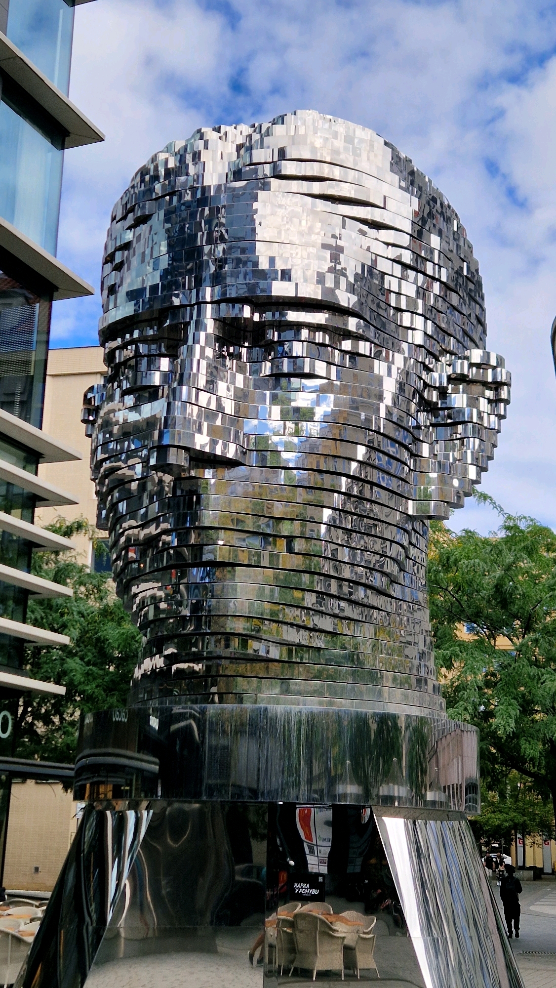 The image displays a large, metallic sculpture of a human head composed of stacked, reflective segments. The sculpture is located in Prague, Czechia. In the background, there are modern buildings with glass facades and trees. The sculpture's reflective surface mirrors the surroundings, including the sky with clouds, the buildings, and some of the trees. The base of the sculpture is also metallic and highly reflective. Reflected in the base are tables and chairs, suggesting an outdoor seating area, and a sign with Czech text reading "KAРФА V POHYBU" which translates to "Kafka in Motion." A person is visible walking on the right side of the image. The lighting suggests it is daytime.
