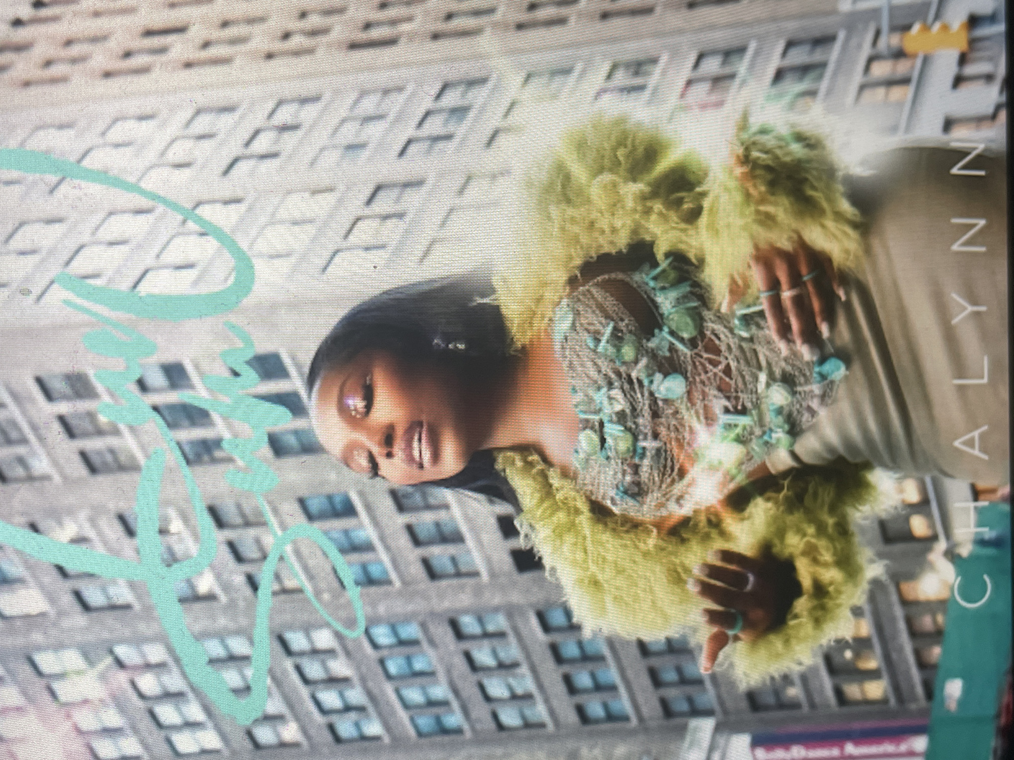 An upright portrait-oriented image features a woman posing against an urban cityscape background, likely a promotional or fashion shot. The central subject is a Black woman with dark hair, looking slightly upwards and to her right, with a serious expression and slightly parted lips. She wears a striking outfit, including a voluminous, light green, fluffy, or feathered jacket/shrug. Beneath it, an intricate, open-weave top with turquoise or light blue stone-like embellishments covers her chest. Her left arm and hand, adorned with several rings, are visible.
The background consists of a tall, grey building with numerous rectangular windows, creating a grid-like pattern typical of a modern city. The lighting suggests daytime. On the right side of the image, running vertically, is the text "CHALYNN" in white capital letters. On the left side, a stylized graphic or partial text, in a light blue or teal color, is visible, featuring swirling lines and curves, possibly part of a logo or another word. The overall aesthetic is glamorous and stylish, set against a generic urban backdrop.