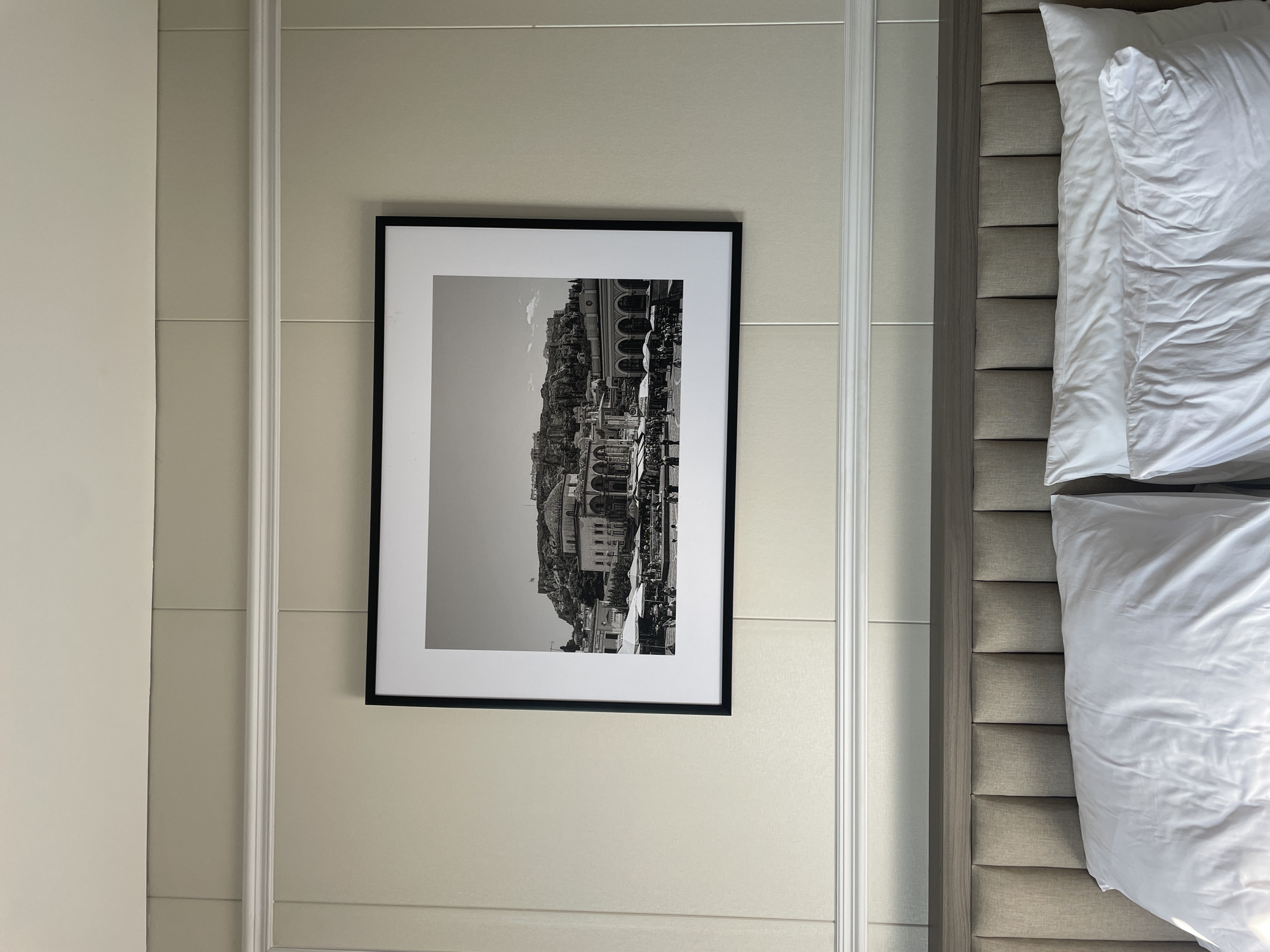 This image depicts a bedroom located in the city of Kayuambon, Indonesia. The primary focal point is a large, horizontally oriented black-framed photograph hanging centrally on a paneled wall painted in a light beige color. A white decorative molding runs horizontally across the wall beneath the photograph, separating the wall into upper and lower sections. The ceiling above is white.
The framed photograph, presented in black and white with a wide white mat, captures a panoramic view. In the background, a prominent rocky hill is visible, topped with ancient architectural ruins, specifically the Acropolis of Athens, Greece, with a flag visible near its summit. Below the Acropolis, the mid-ground and foreground of the photograph show a bustling public square, identifiable as Monastiraki Square. This square is densely populated with numerous individuals, market stalls, and outdoor seating arrangements, many shaded by various types of umbrellas. Several multi-story buildings with arched ground-level facades line the square.
Below the photograph, the upper portion of a bed is visible. It features a modern headboard upholstered in a light, neutral-toned fabric, designed with distinct vertical padded panels. This headboard is capped with a dark wood or wood-look trim piece. On the bed, two white pillows with white pillowcases are present, occupying the lower segment of the frame.