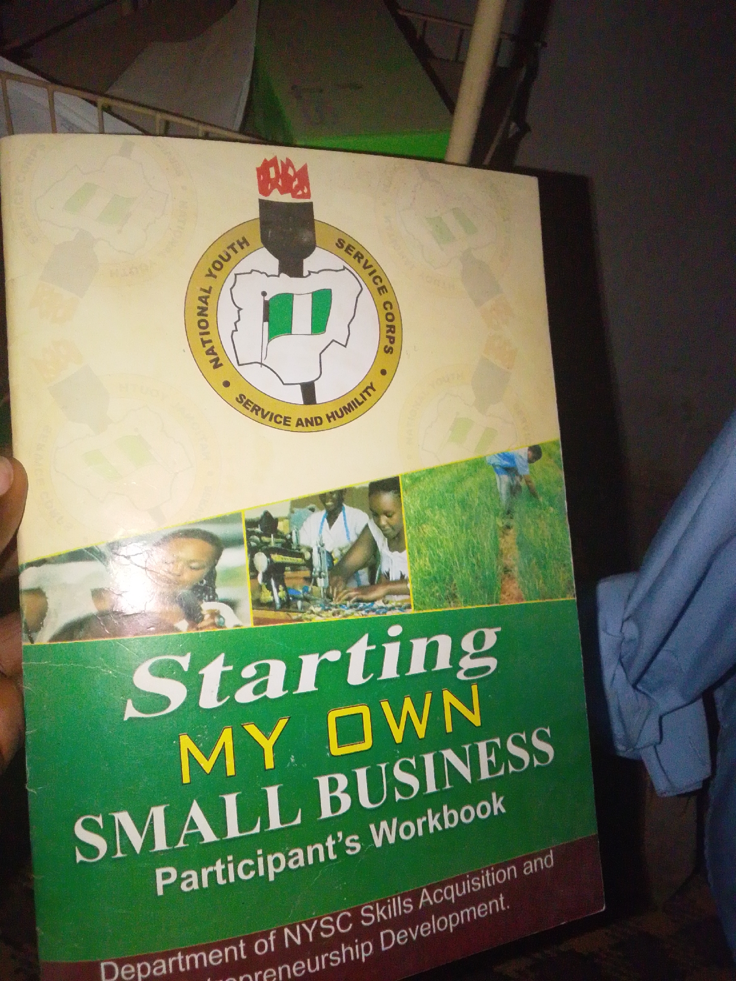 The cover of a workbook titled "Starting My Own Small Business" is displayed. The book features the National Youth Service Corps (NYSC) logo prominently at the top, depicting the Nigerian flag within a shield. Below the logo, three inset images showcase activities related to business and skill development: a woman working with electronics, individuals in a workshop setting, and a person tending to a farm. The workbook is presented as a participant's guide for skills acquisition and entrepreneurship development.