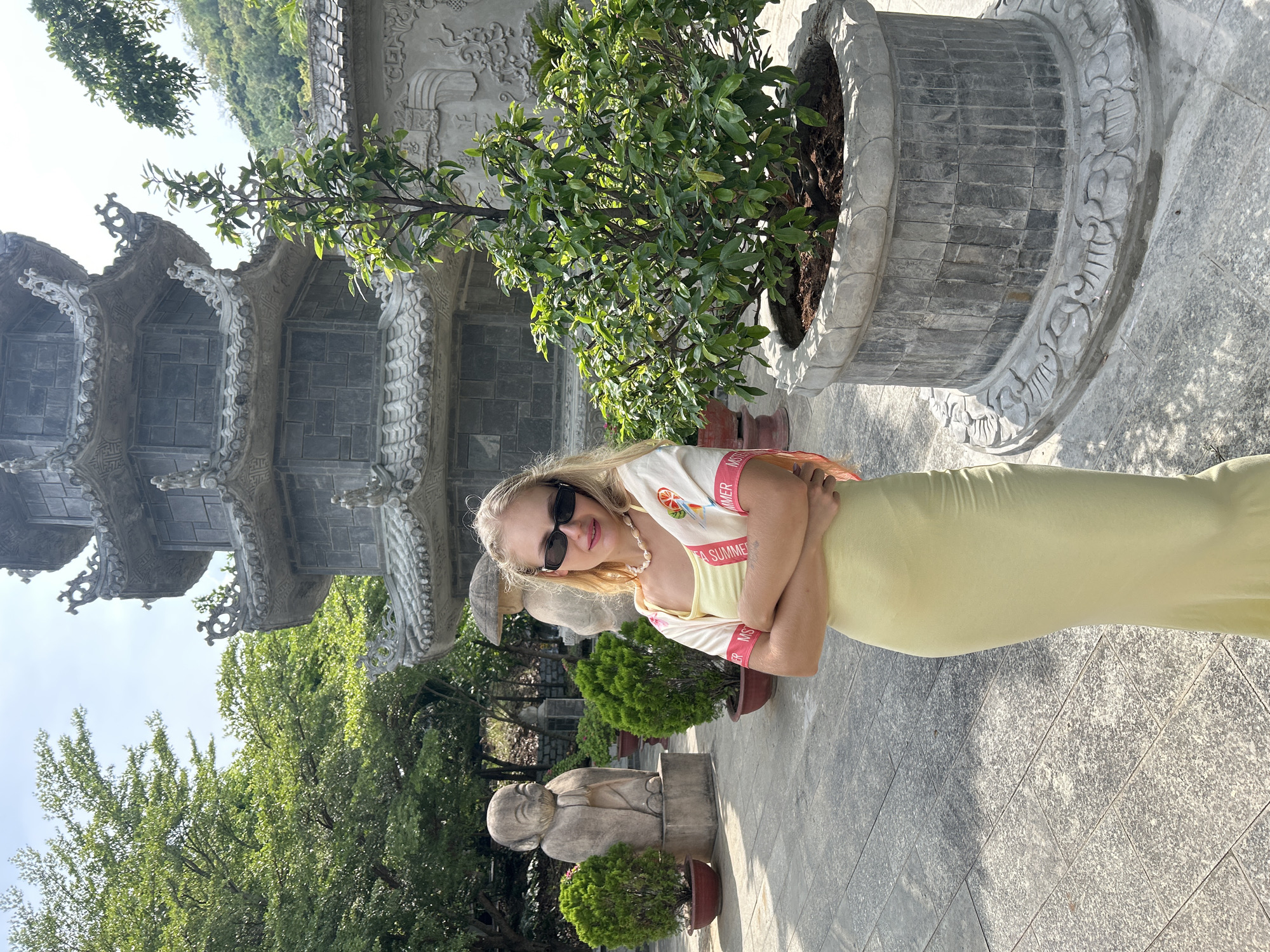 A blonde woman stands in front of a multi-tiered pagoda in Nam Nha Trang Ward, Vietnam. She is wearing a pale yellow slip dress and a cropped light-colored cardigan with pink trim. She has on black-rimmed sunglasses and a shell necklace. Her arms are crossed, and she is looking at the camera. The pagoda is made of grey stone and is surrounded by trees and potted plants. To the left of the woman are two stone statues, one of a seated figure and another of a standing figure with a conical hat. A large potted tree with green leaves is to the woman's right. The ground is paved with grey stone tiles. The sky is bright and sunny.