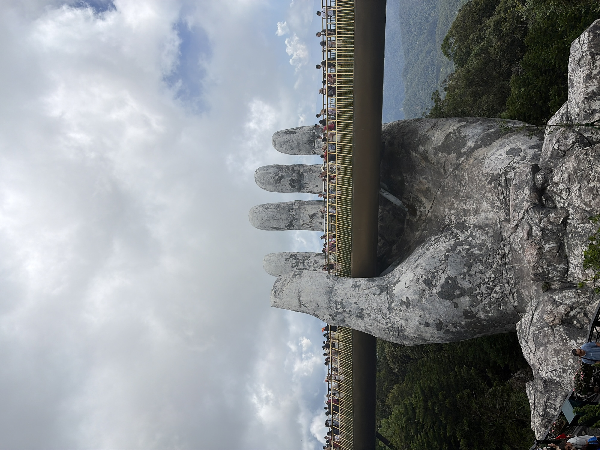 The image shows the Golden Bridge in Đà Nẵng, Vietnam, supported by two giant stone hands. A crowd of people are on the bridge. Lush green vegetation covers the surrounding hills. The sky is overcast with scattered clouds.