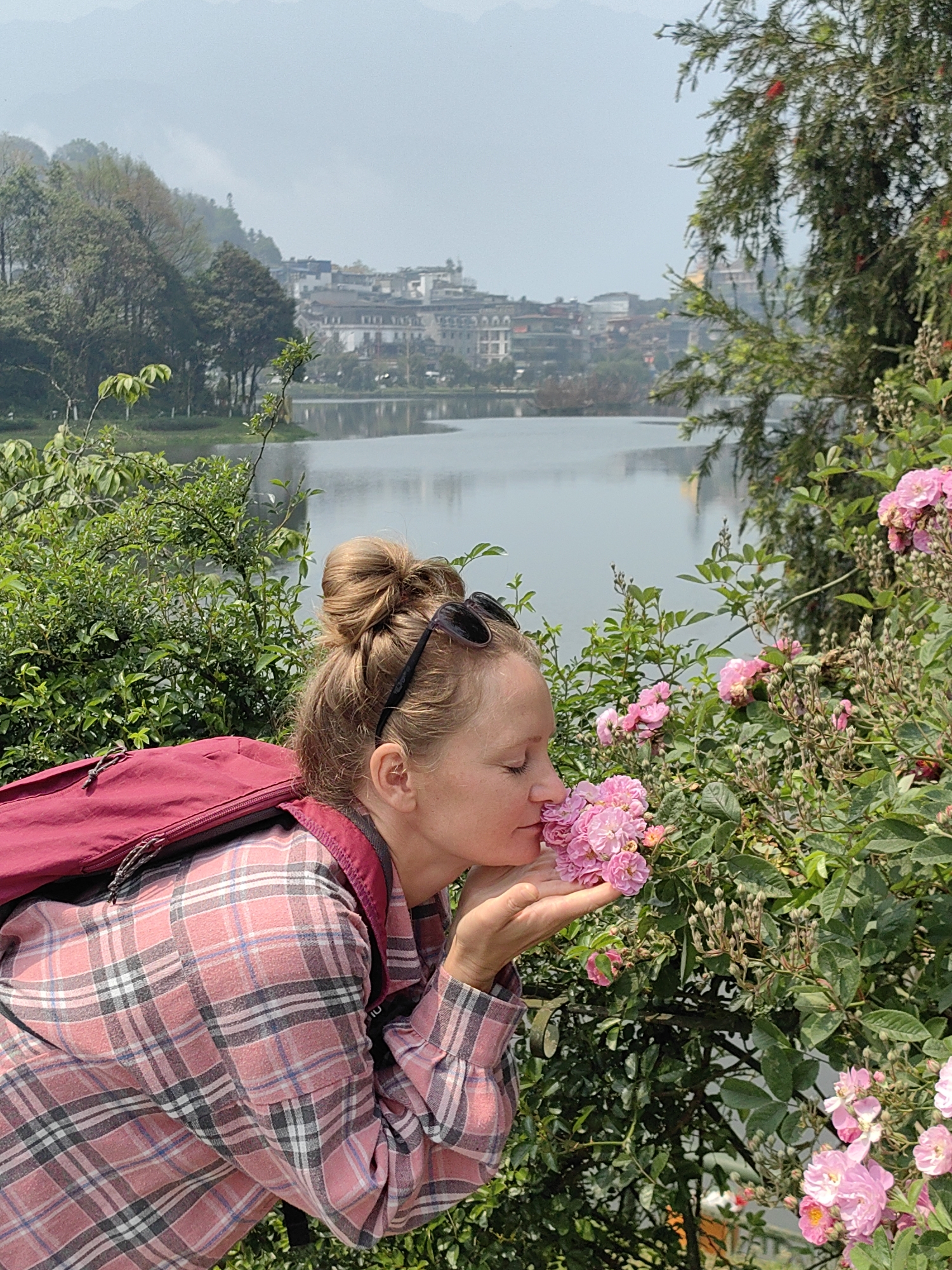 An adult female is depicted in the foreground, leaning towards a cluster of small pink roses, with her eyes closed as she smells them. Her left hand is cupped beneath the flowers. She has light brown hair styled in a high bun, and dark sunglasses are resting on top of her head. She is wearing a red and black plaid long-sleeved shirt and a maroon backpack. The immediate surroundings consist of dense green foliage and flowering bushes. In the background, a calm body of water, identifiable as Sa Pa Lake in Sa Pa, Vietnam, reflects the sky and structures. Numerous multi-story buildings of Sa Pa line the far bank of the lake, with hazy mountains visible beyond the cityscape under a light blue-grey sky.