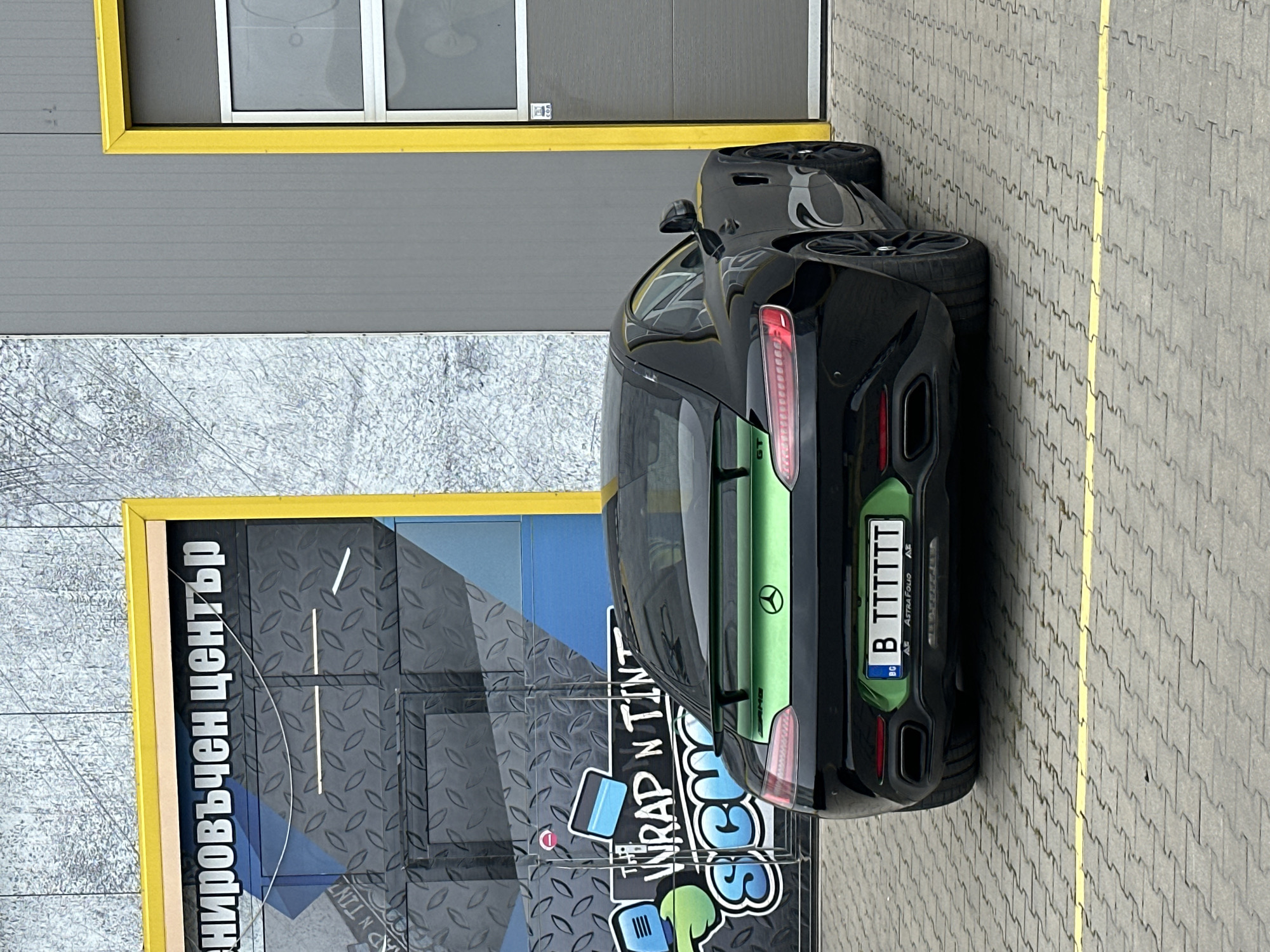 A black Mercedes-AMG GT sports car is parked outdoors, angled with its rear facing the viewer, against a building in Varna, Bulgaria. The car features a distinctive green stripe running along its trunk lid and rear spoiler. Its rear taillights are visible, and the car's side profile shows a sleek design.
The Bulgarian license plate reads "B TTTTTT", with "B" indicating the Varna region. The license plate frame displays "BG", "AF", and "ASTRA FOLIO". Small "AMG" and "GT" badges are partially visible on the car's rear.
The setting appears to be outside a commercial establishment, possibly a car wrapping or tinting shop. The background includes a building with gray horizontal siding and a large, textured gray door or panel. This large panel has a prominent blue section and is framed by yellow trim. White Cyrillic text "нировъчен център" (likely part of "Бронировъчен център" - "Wrapping/Armoring Center") is visible on the building. A colorful logo below it includes the text "WRAP N TINT" and "SCH", indicating the business's services.
The ground is paved with interlocking gray pavers, and a yellow line runs across the pavement to the right of the car. The lighting suggests an overcast or generally bright daytime environment. The image is oriented sideways, requiring rotation to view naturally.