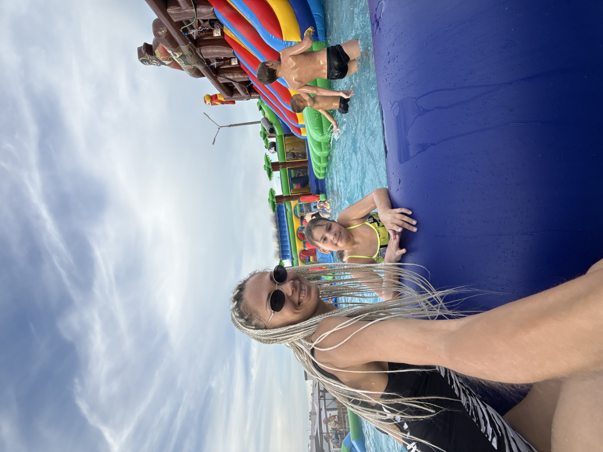 In this selfie taken at a water park in Rusanovo, Russia, a woman with long blonde braids and sunglasses smiles at the camera. She is wearing a black swimsuit. Behind her, a young girl in a yellow bikini is also in the water, smiling. In the background, a large inflatable water slide structure is visible, with children playing on it. The water is blue and clear, and the sky is cloudy. The overall mood is one of fun and enjoyment at a water park on a cloudy day.