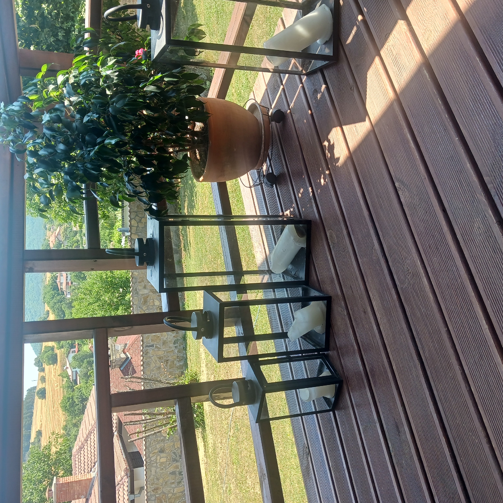 The image shows a wooden deck with four lantern-style candle holders and a potted plant. The deck is made of dark brown wooden planks. The lantern holders are made of black metal with glass panels. Each holder contains a white candle, some of which appear to be partially burned. A large green plant in a terracotta pot sits on a wheeled stand to the right of the lanterns. The background shows a village with houses, trees, and rolling hills under a clear sky. The village of Sulitsa, Bulgaria is depicted in the background.