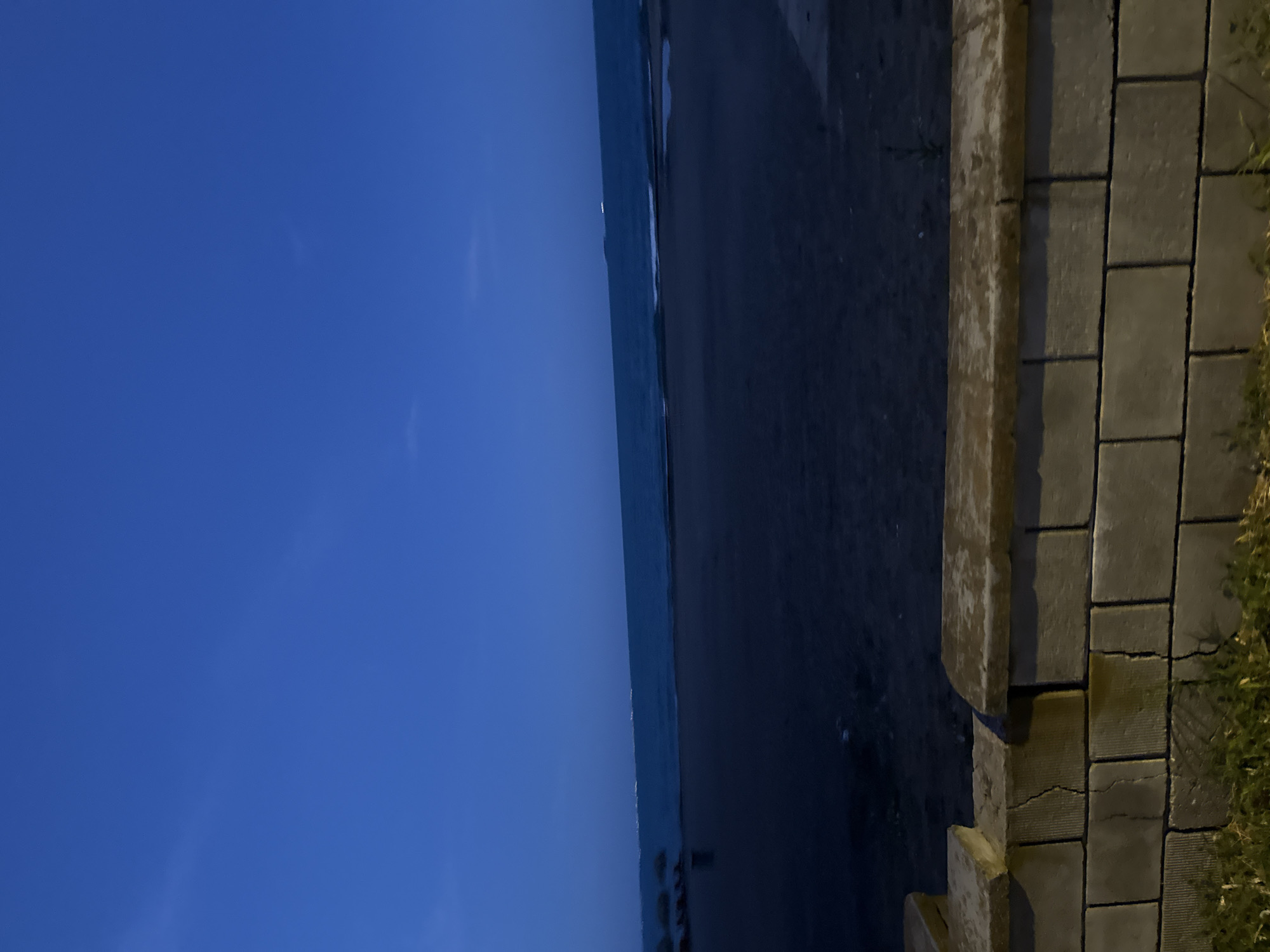The image depicts a dark, twilight scene at the Burgas beach in Burgas, Bulgaria. In the foreground, a low wall constructed of grey concrete blocks topped with weathered concrete slabs runs horizontally across the frame. Some of the blocks show cracks and staining. Green grass and weeds grow at the base of the wall.
Beyond the wall, a dark sandy beach stretches into the mid-ground. The sea is visible in the background, its surface appearing dark blue and calm, with small waves lapping at the shore. In the far distance, on the horizon, a faint line of lights suggests the presence of distant buildings or a cityscape. The sky above is a deep, clear blue, indicative of the late evening or early morning hours. No people or specific activities are clearly discernible due to the low light conditions.